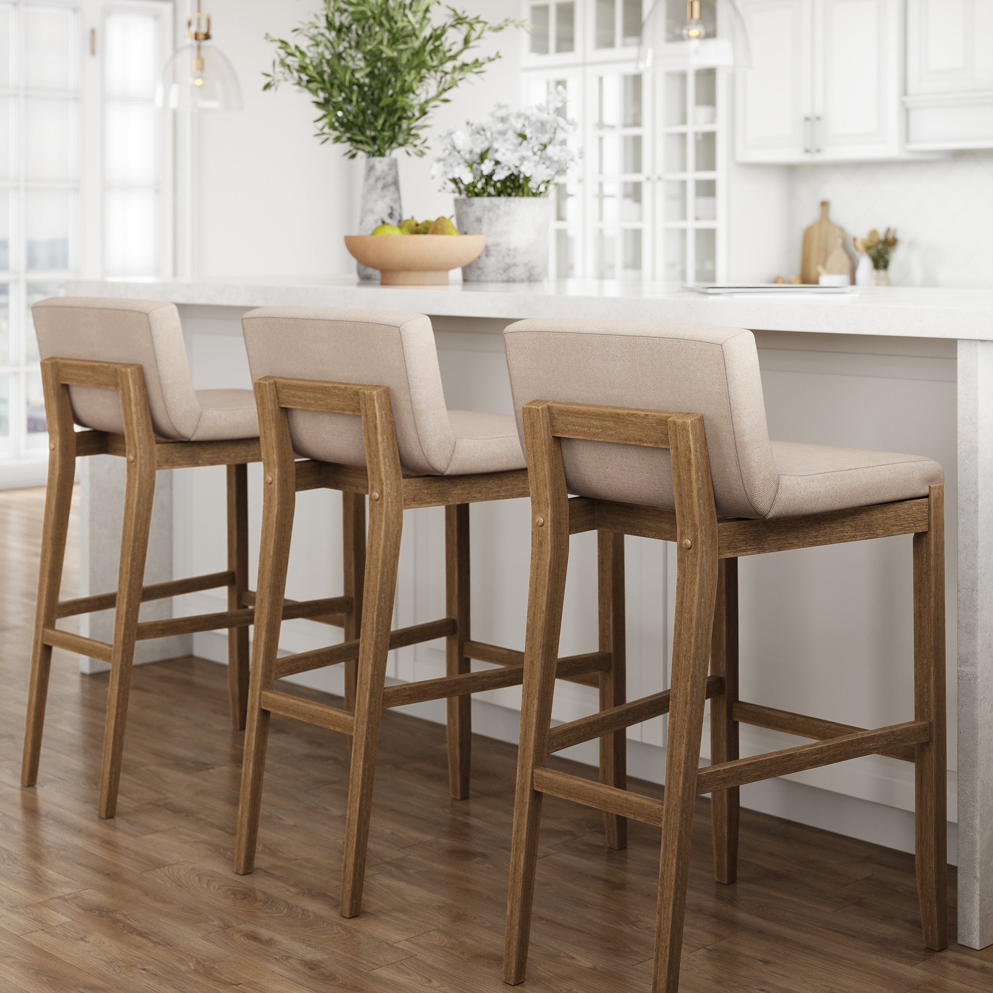 Three wooden barstools with beige upholstered seats are lined up at a white kitchen island in a bright, modern kitchen with wooden floors and decorative plants on the counter.