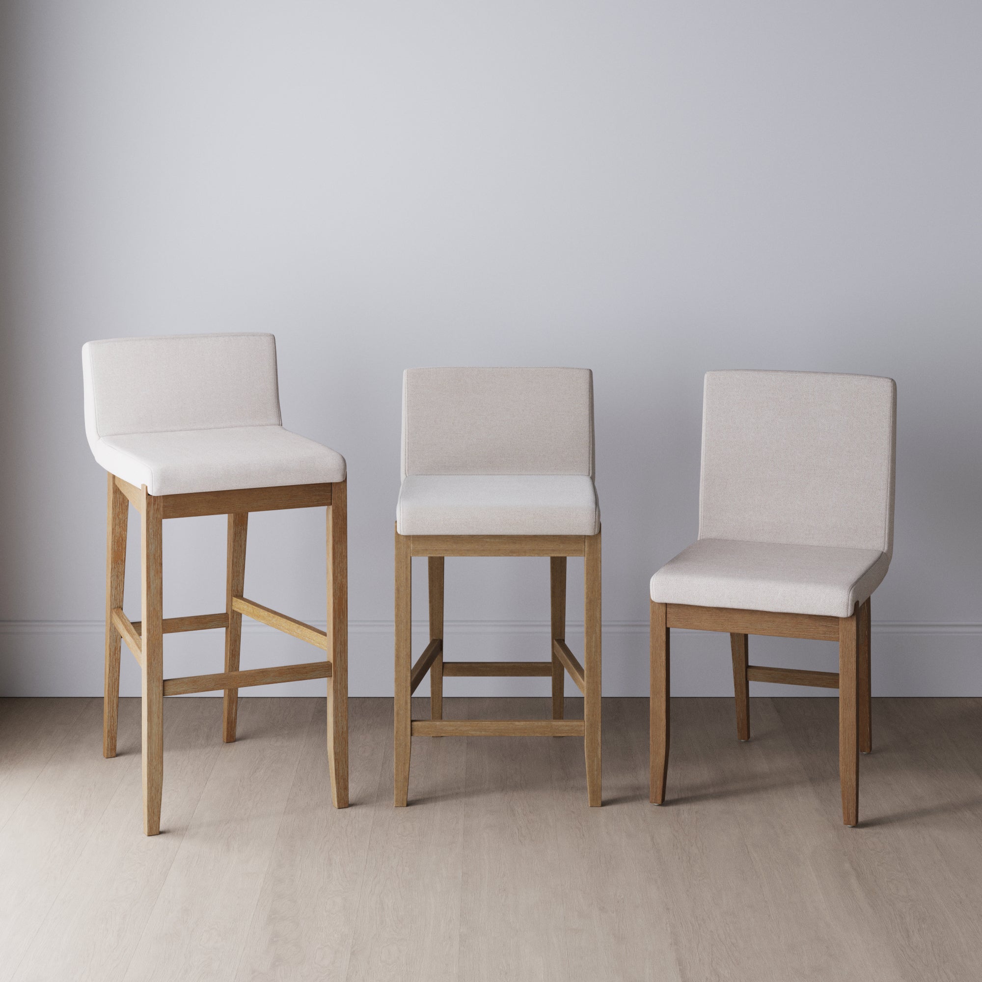 Three wooden chairs with light beige upholstered seats and backs, each at different heights, are arranged side by side against a plain light gray wall on a light wood floor.