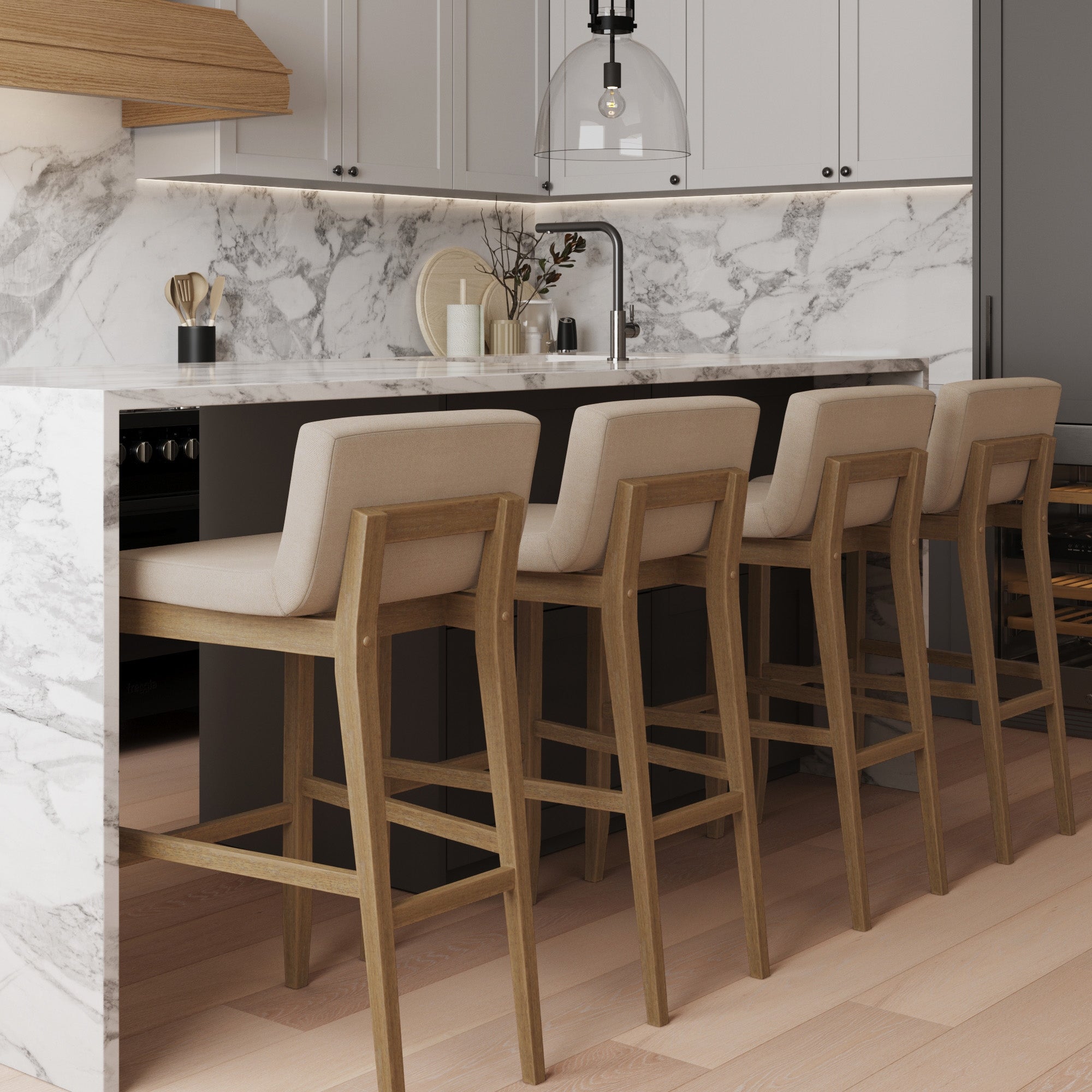 A modern kitchen features a marble island with four wooden bar stools, beige cushions, light wood flooring, white cabinets, and minimalist decor including vases and candles near the sink.