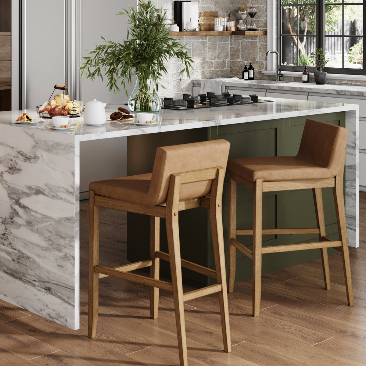 Two tan upholstered bar stools sit at a marble kitchen island with a bowl of fruit and a vase of greenery.