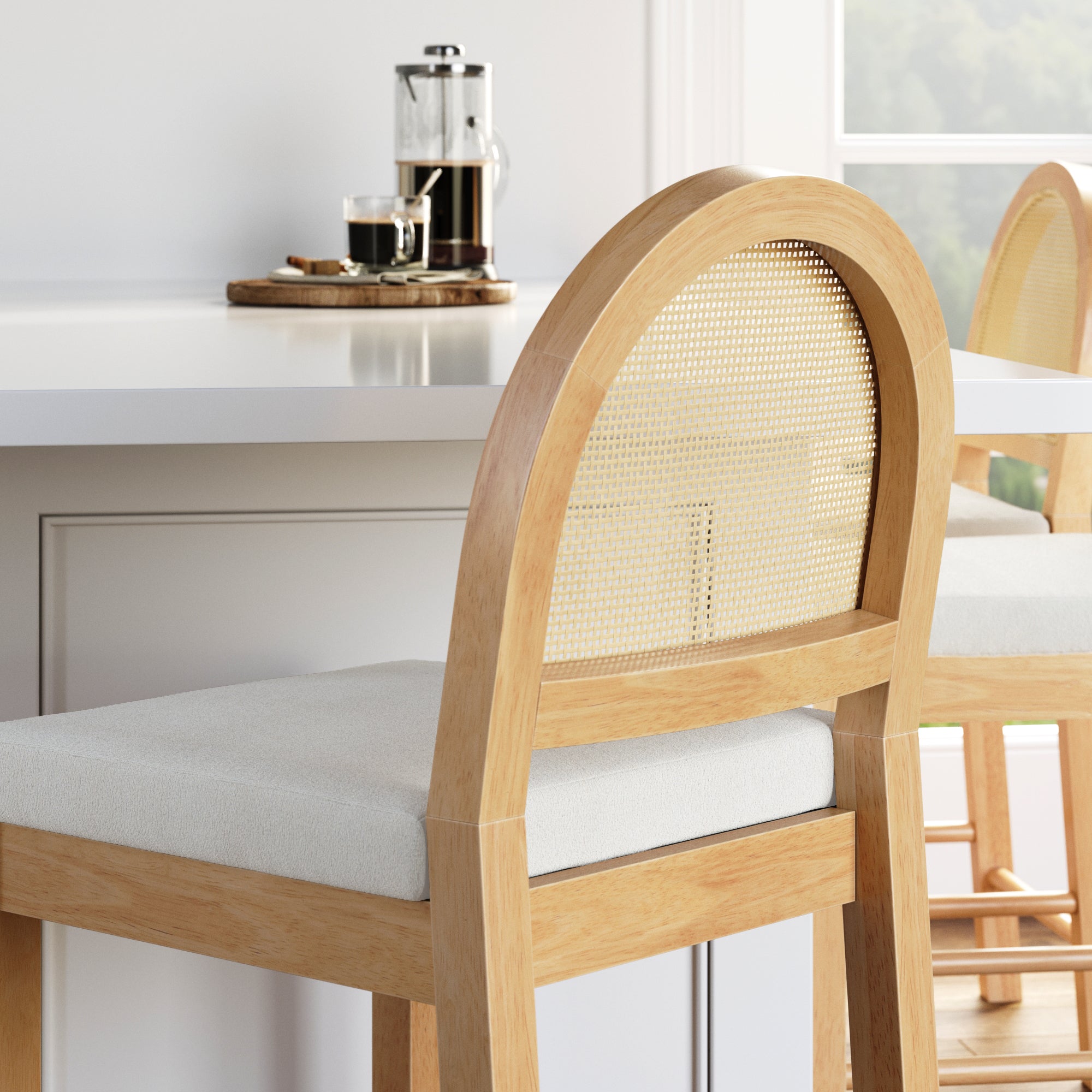 A light wood chair with a woven backrest and white cushion sits by a white kitchen counter. A French press and cup are on a tray in the background near a window with greenery outside.