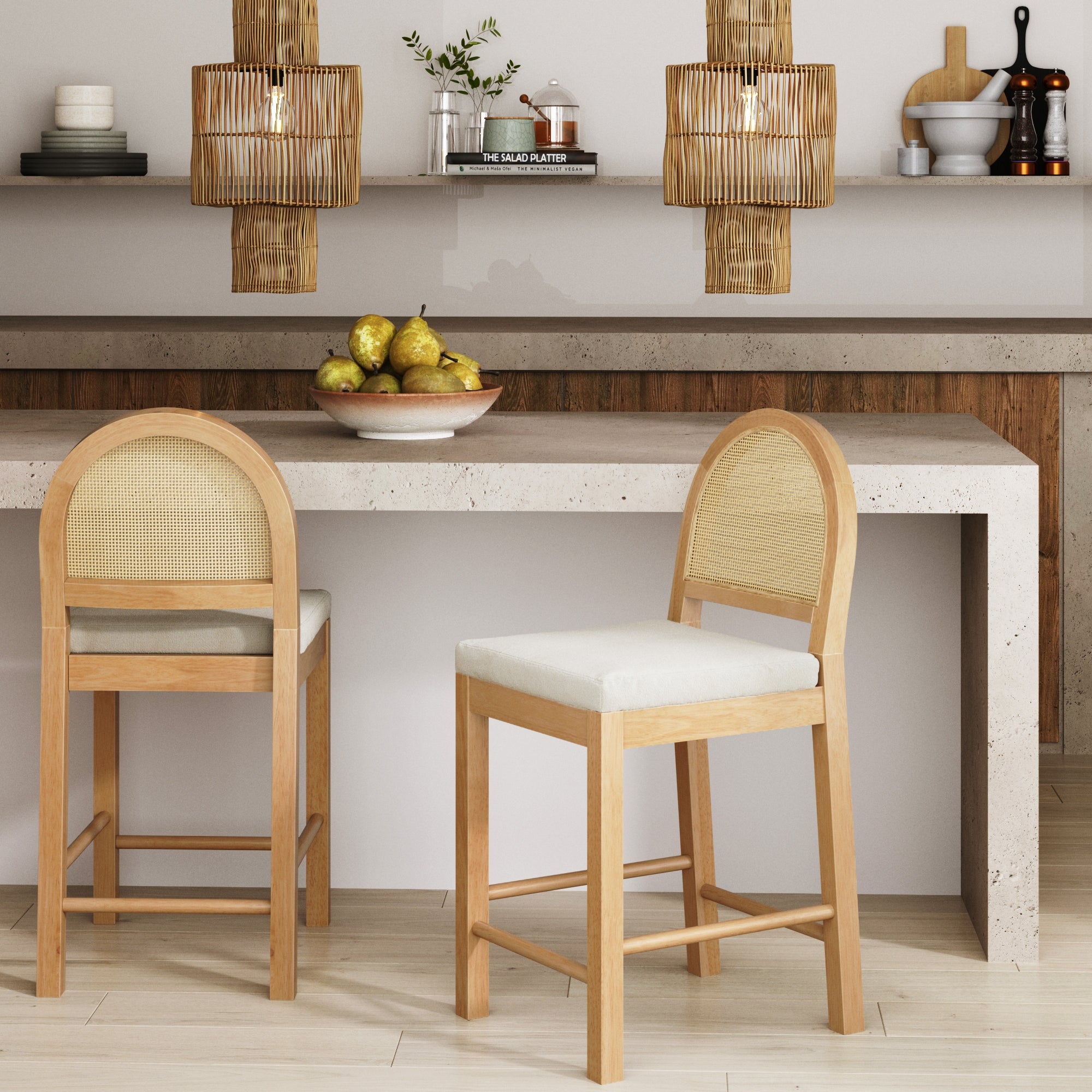 A modern kitchen features a stone island with two wooden barstools, a bowl of pears, wicker pendant lights, and shelves with plants, books, and dishware against a white and wood-accented wall.