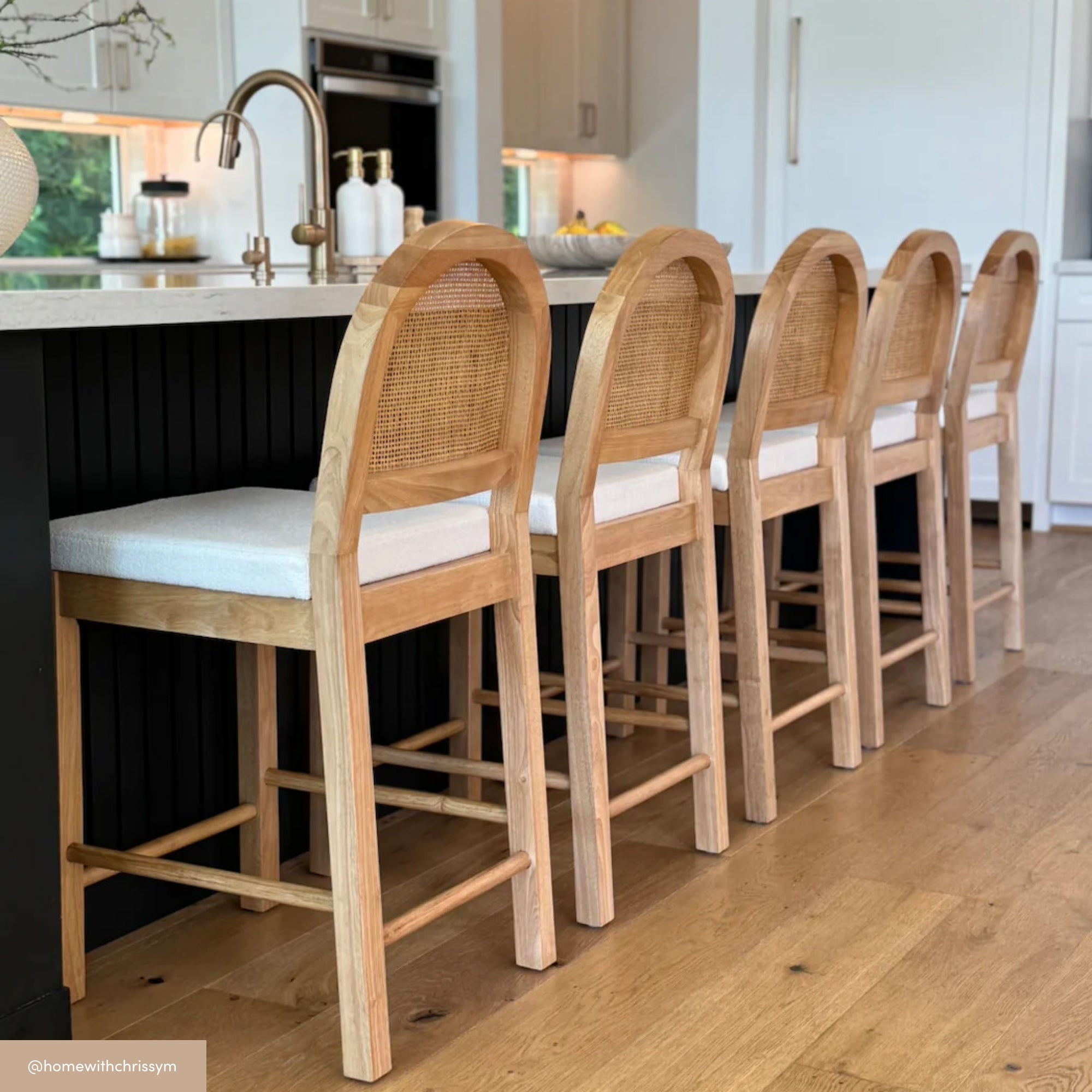 Four wooden bar stools with cane backs and white cushions are lined up along a modern kitchen island with a sink and faucet. The kitchen has light wood flooring and white cabinetry.