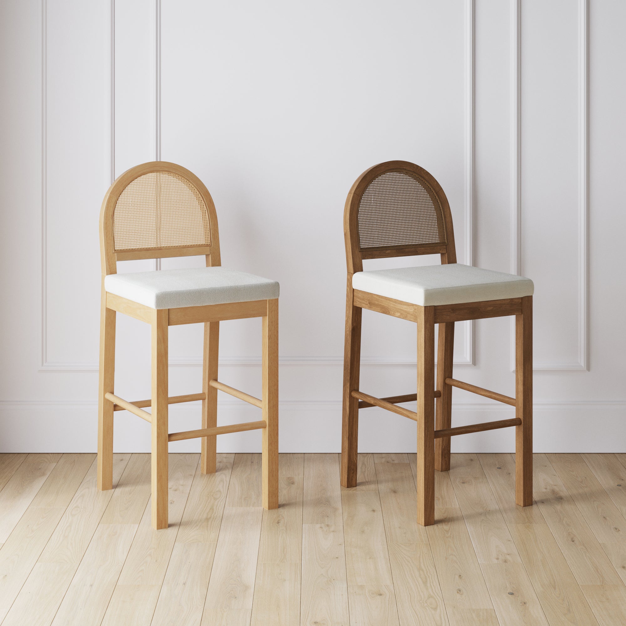 Two wooden bar stools with light-colored cushioned seats and rounded, woven backs are placed side by side on a light wood floor against a white paneled wall.