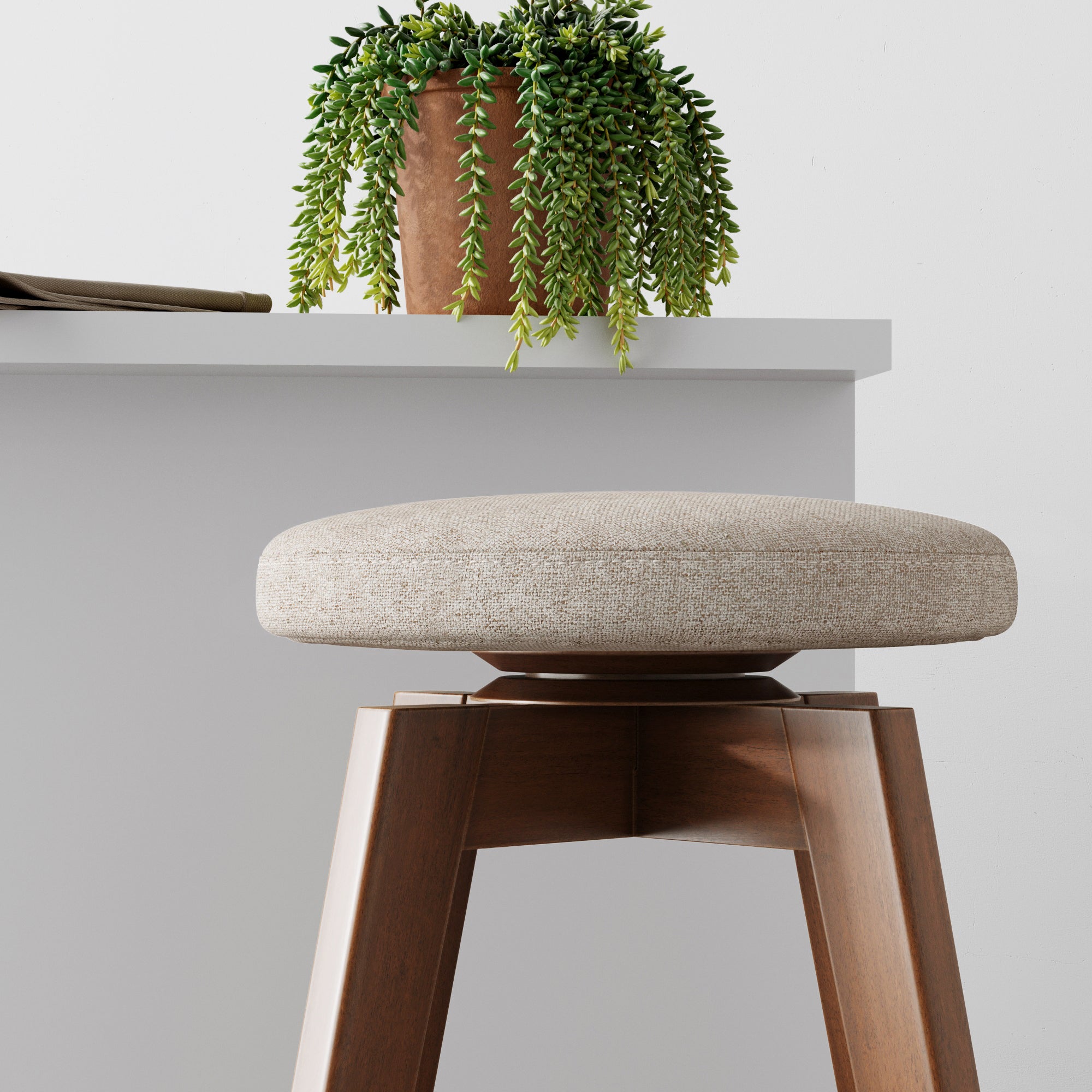 A modern wooden stool with a beige cushioned seat stands in front of a white surface topped with a potted trailing plant and a closed book. The background is minimal and bright.