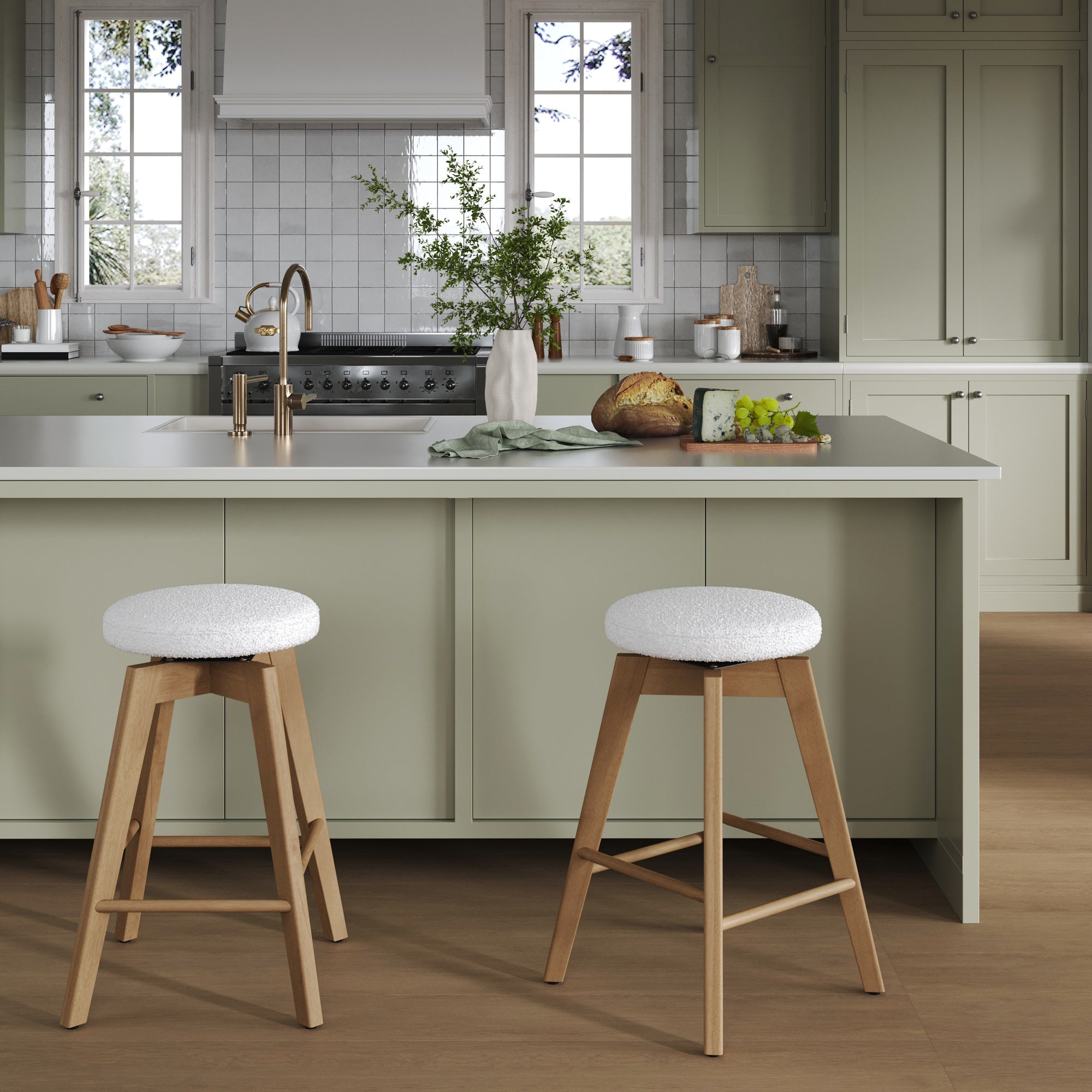 A modern kitchen with light green cabinets, a central island with a white countertop, two wooden bar stools with white cushions, and various kitchen items and plants neatly arranged. Natural light streams through two windows.
