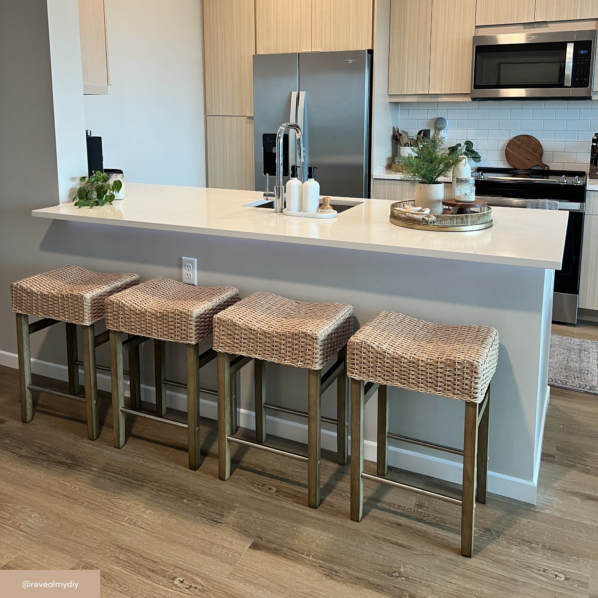 A modern kitchen with a white island countertop, four woven bar stools, stainless steel appliances, light wood cabinets, and decorative plants on the counter.