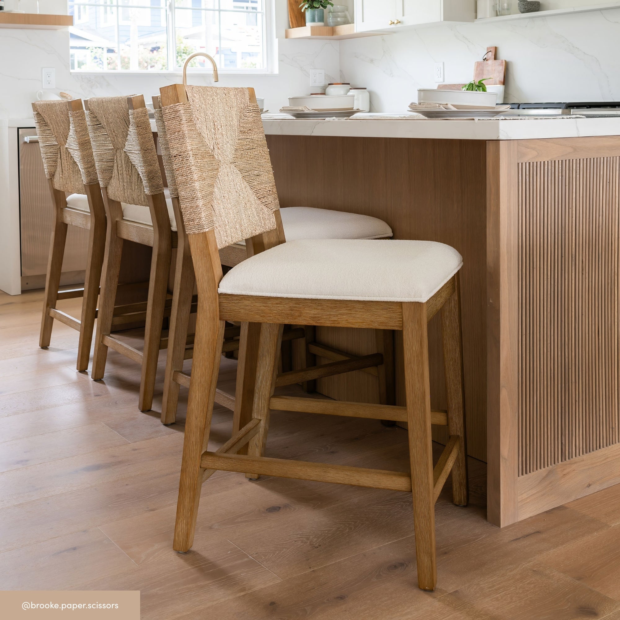 Four wooden bar stools with woven backrests and cream cushions are lined up at a light wood kitchen island in a bright, modern kitchen with wooden floors and white countertops.