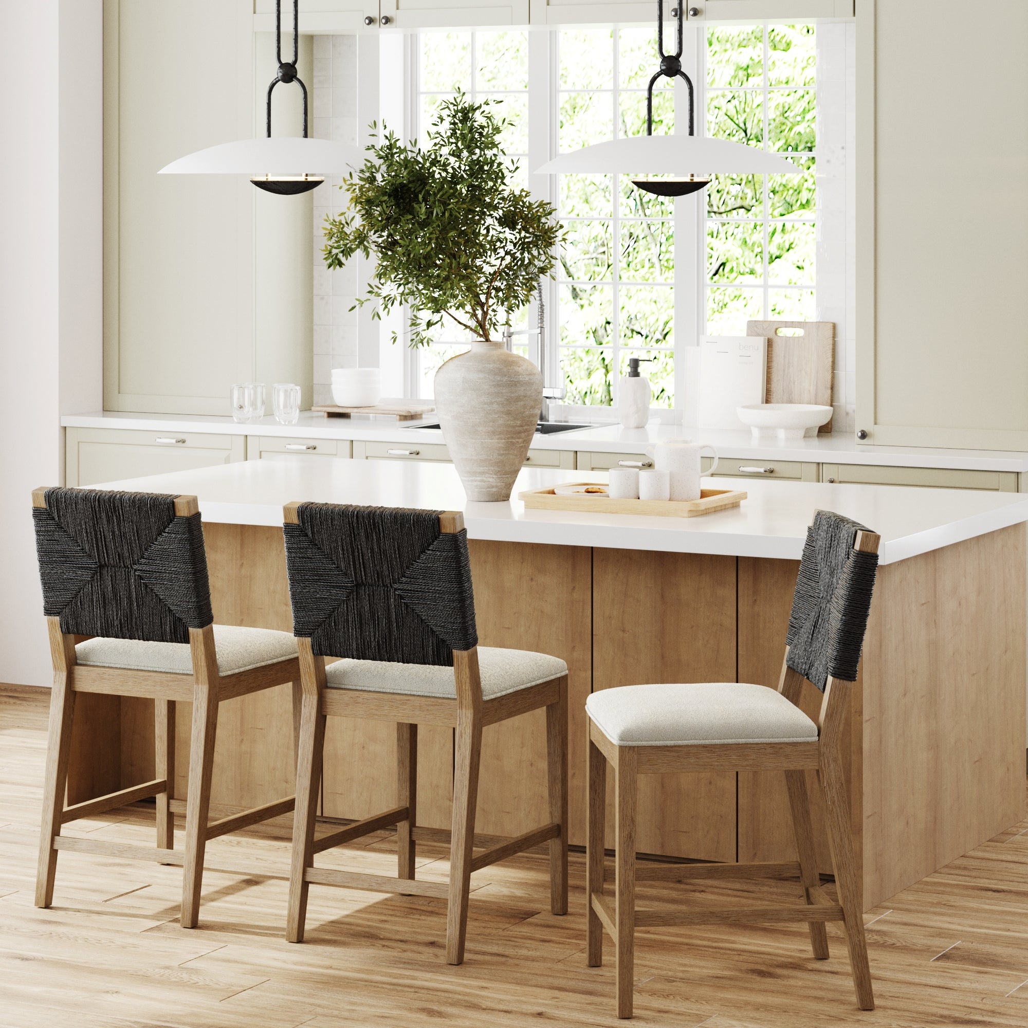 A modern kitchen with light wood flooring, a white island, three wooden bar stools with woven black backs, a large vase with greenery, and large windows letting in natural light.
