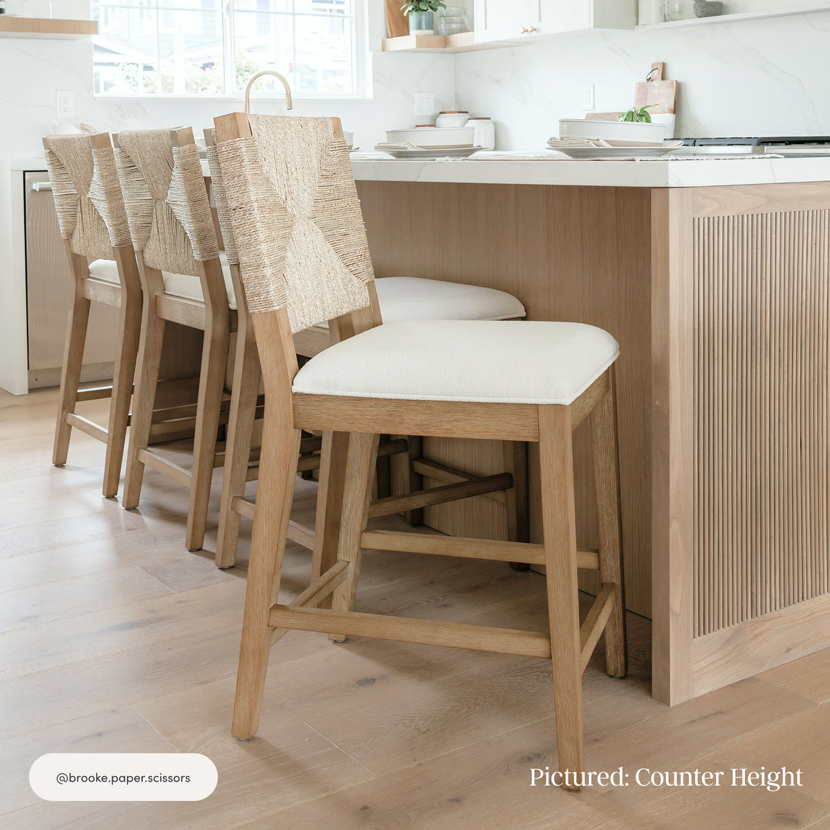 Three Nathan James Boucle & Seagrass Bar Stool Light Brown (Set of 2) 29″ wooden bar stools with cream-colored boucle fabric seats and woven seagrass backrests lined up at a kitchen island.