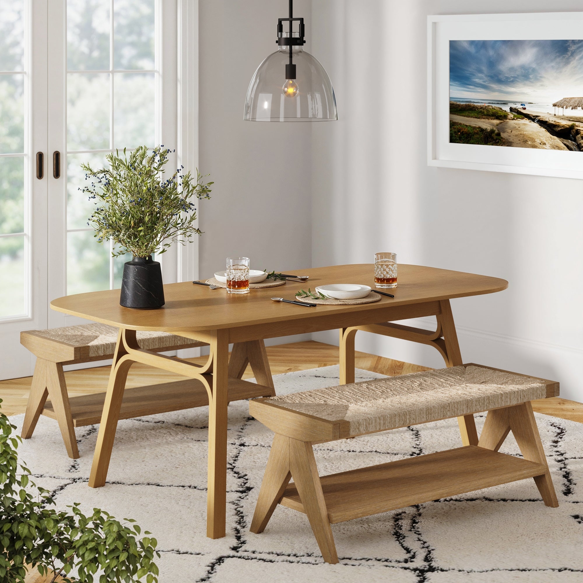 A modern dining room features a wooden table with two matching benches, a black vase with greenery, place settings, glasses with drinks, a pendant light overhead, a framed landscape photo, and large windows in the background.