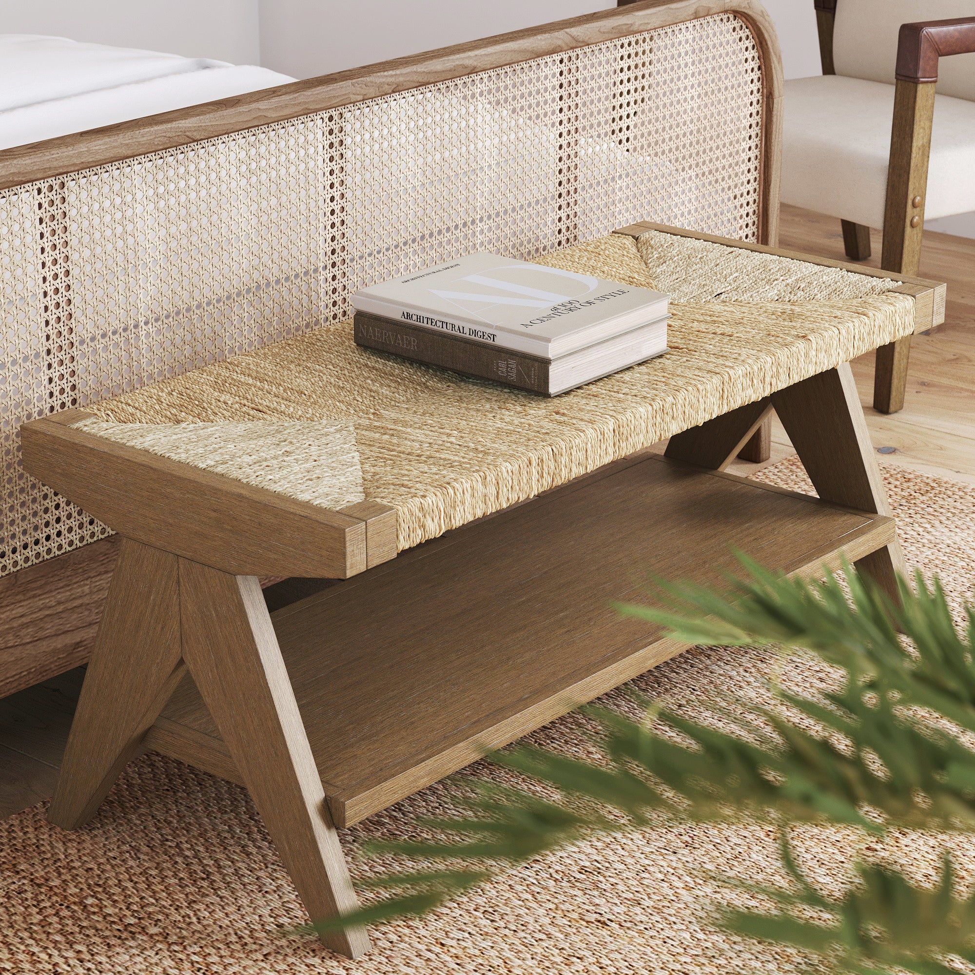 A wooden bench with woven seat top and lower shelf, placed at the foot of a bed with a cane headboard. Two books rest on the bench, and a leafy plant is partially visible in the foreground.