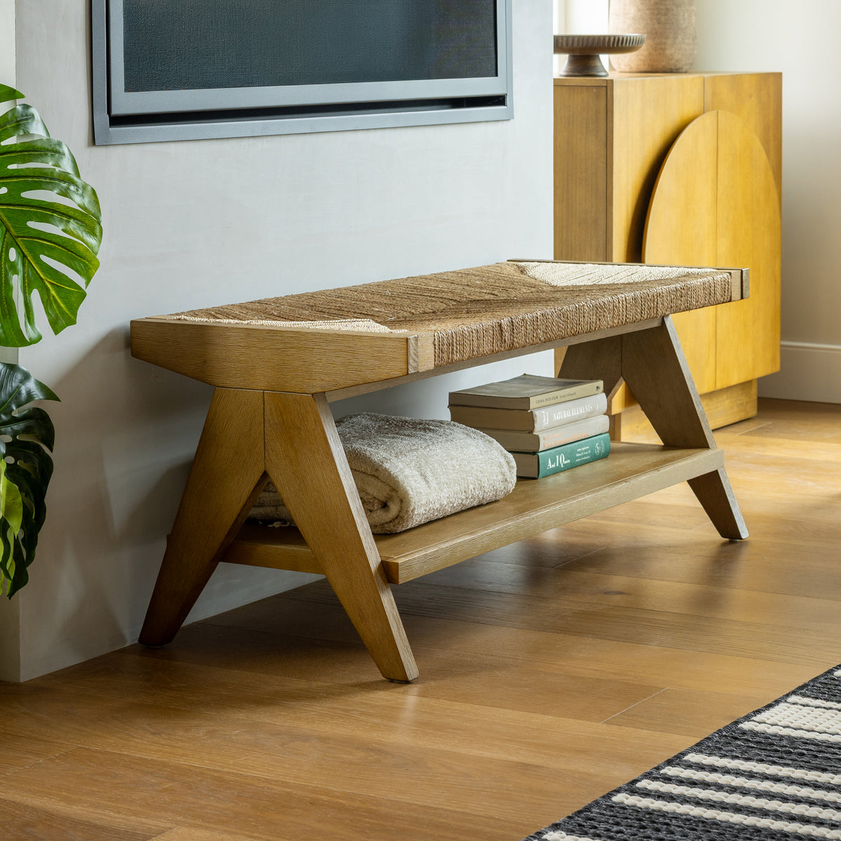 Nathan James Wood & Seagrass Entryway Bench with Shelf featuring a hand-woven seagrass seat, solid wood legs, and a lower shelf holding a folded blanket and books.