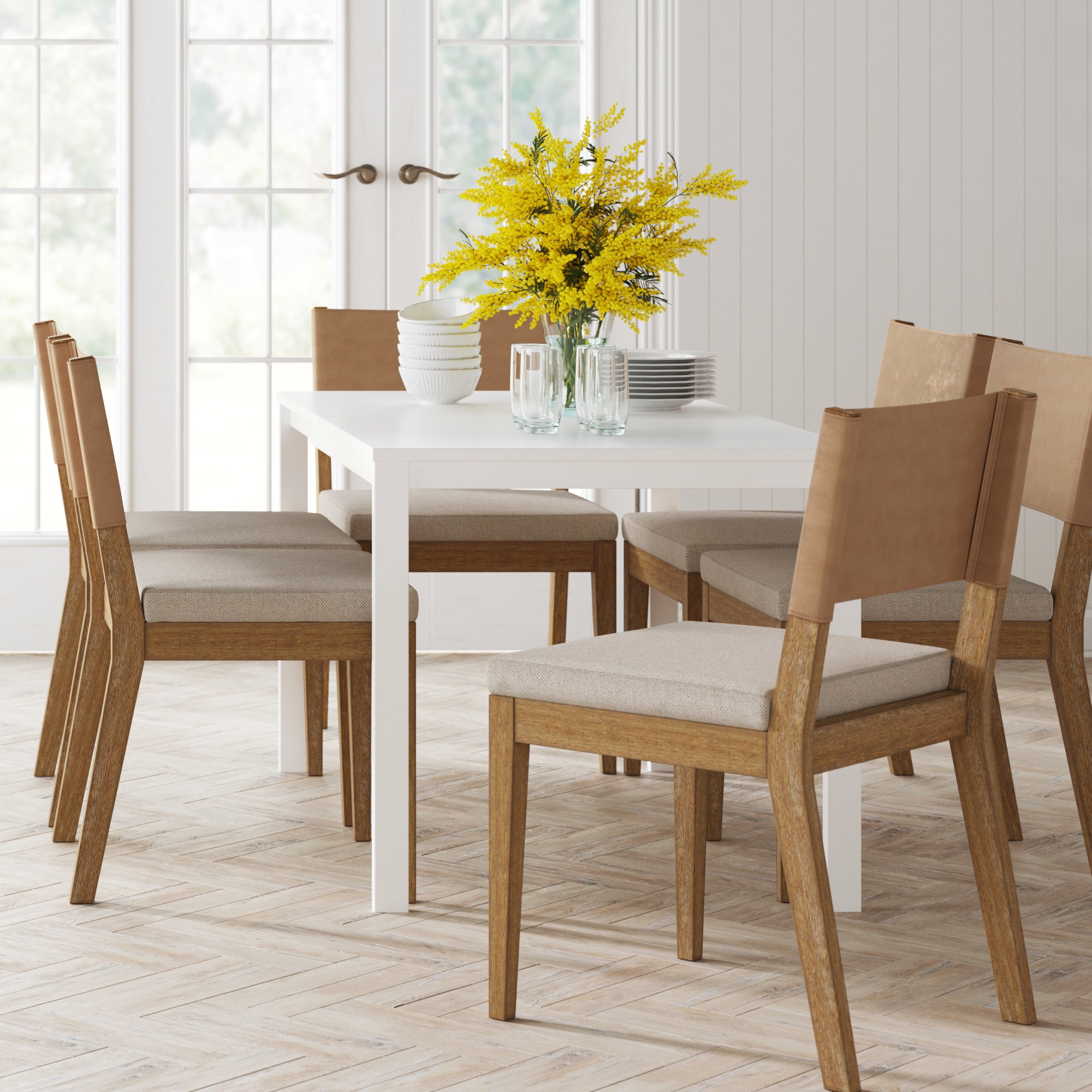 A modern dining room with a white table set for four, surrounded by wooden chairs with tan cushions. A vase with yellow flowers, bowls, plates, and glasses are neatly arranged on the table. Large windows let in natural light.