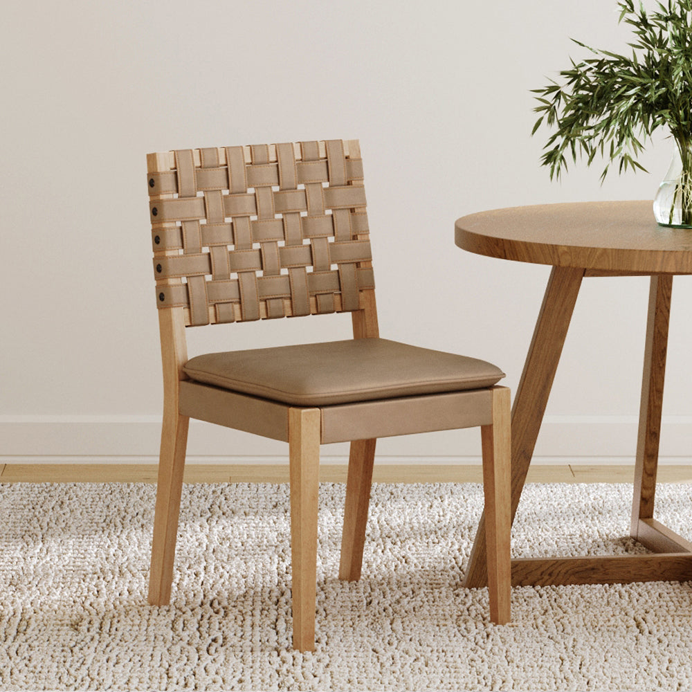 Dining chair with a light wood frame, a woven tan strap backrest, and a solid tan seat cushion, positioned next to a round wooden table.