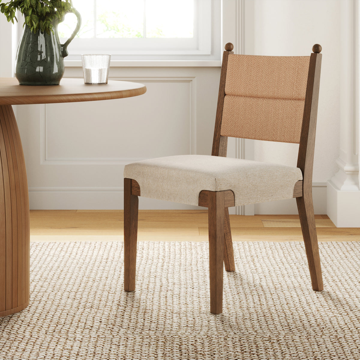 A wooden dining chair with a woven back panel and light-colored upholstered seat next to a round wooden table.