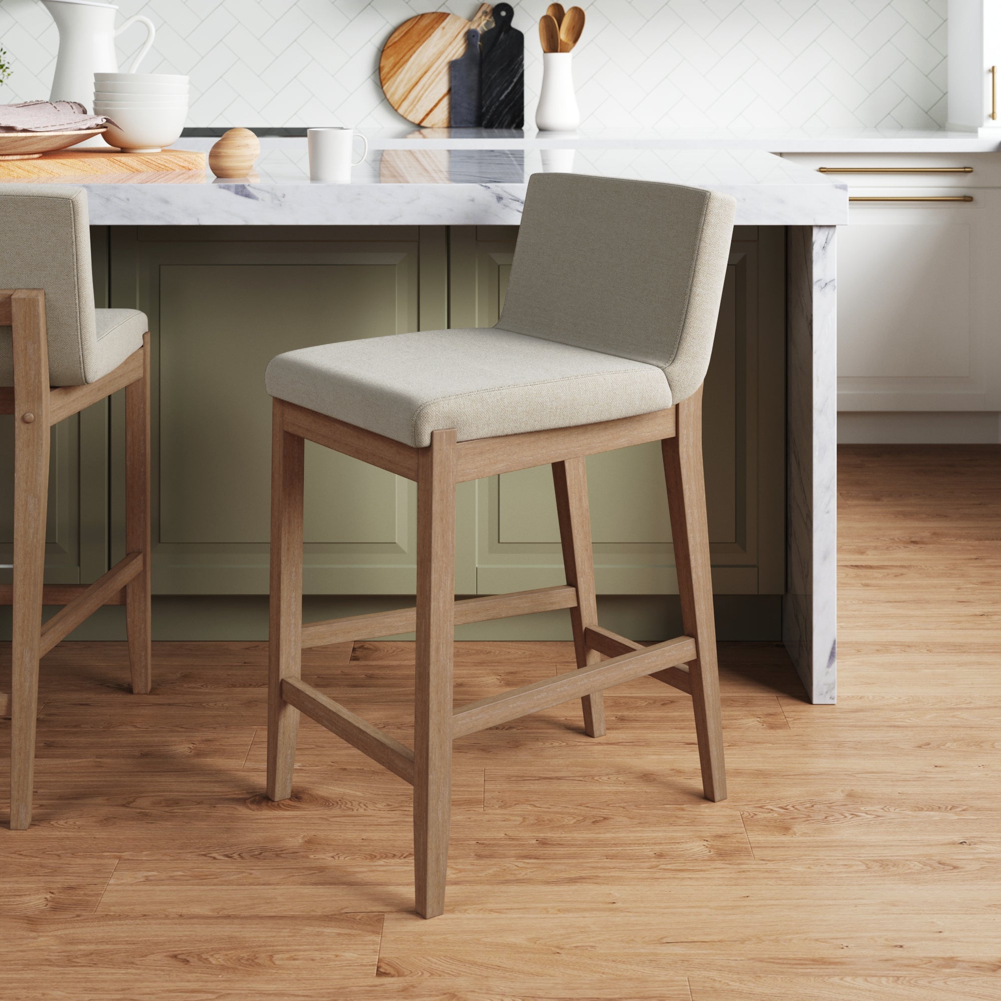 A modern kitchen with a light wood floor features a light gray cushioned bar stool with wooden legs in front of a marble-top island and sage green cabinets. Kitchenware and decor are on the counter in the background.