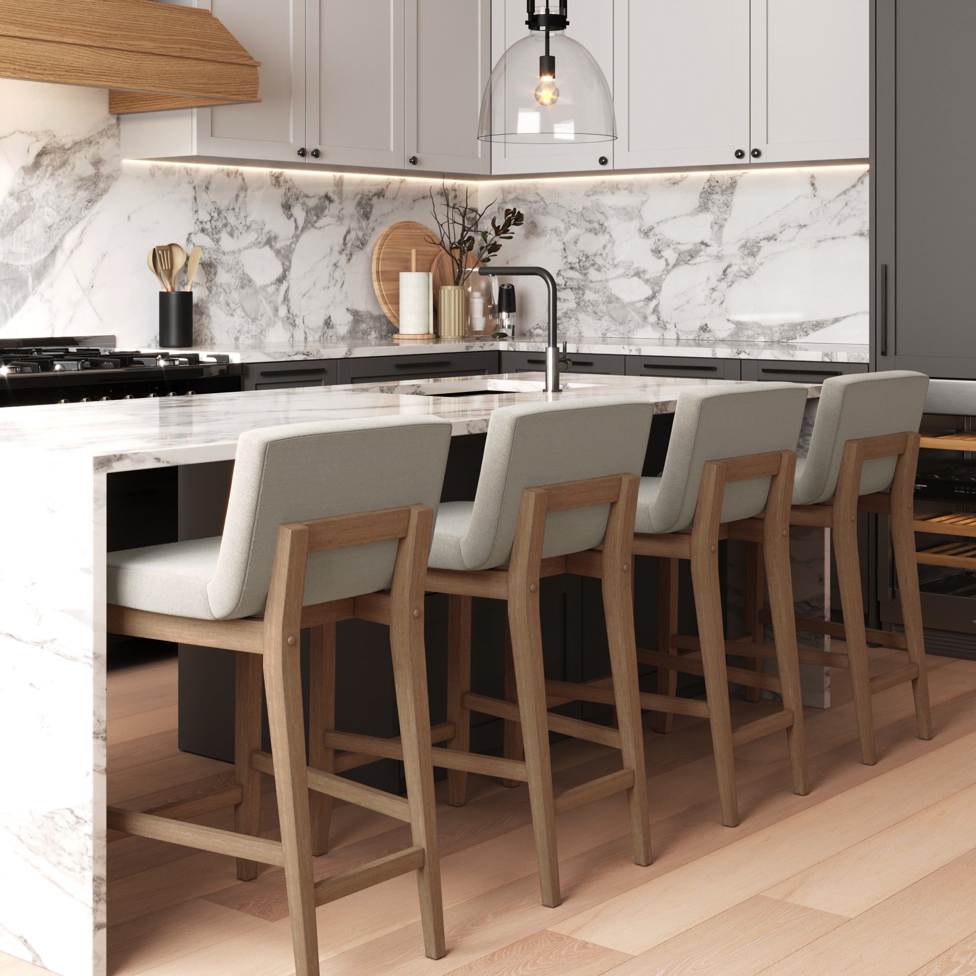 Modern kitchen with a marble island, four wooden bar stools with gray cushions, light wood floors, white cabinets, under-cabinet lighting, and a glass pendant light above the sink.