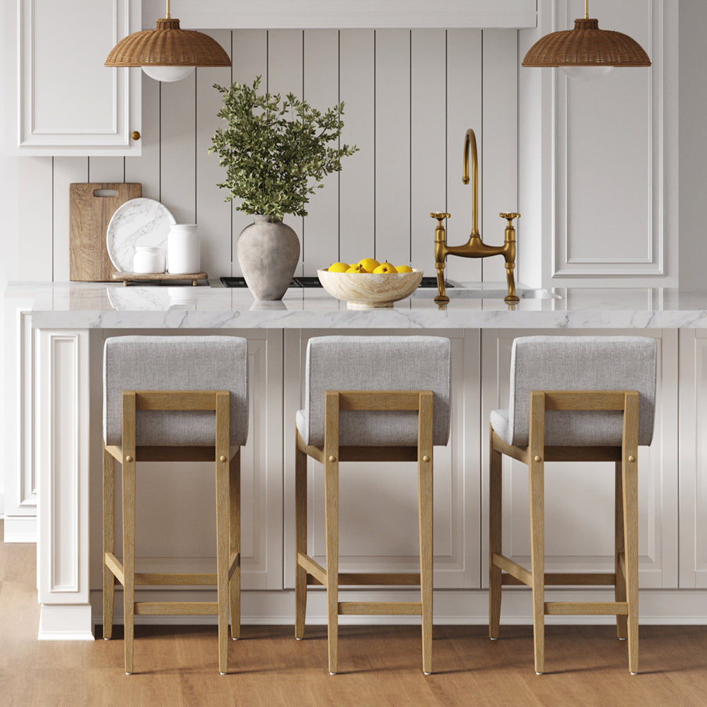 Three gray upholstered bar stools with wooden frames lined up at a marble kitchen island.