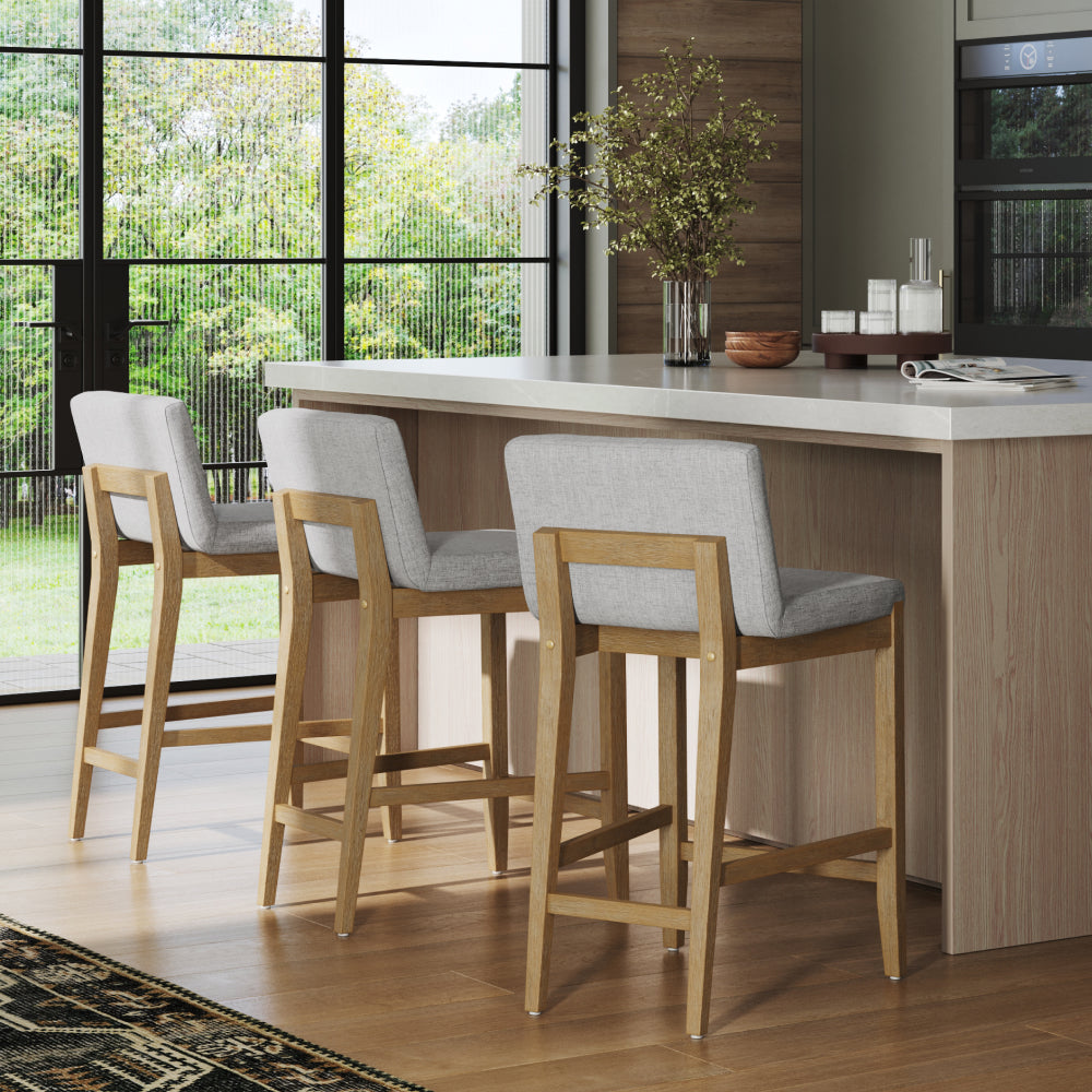 Three light-gray upholstered bar stools with wooden frames lined up at a kitchen island.