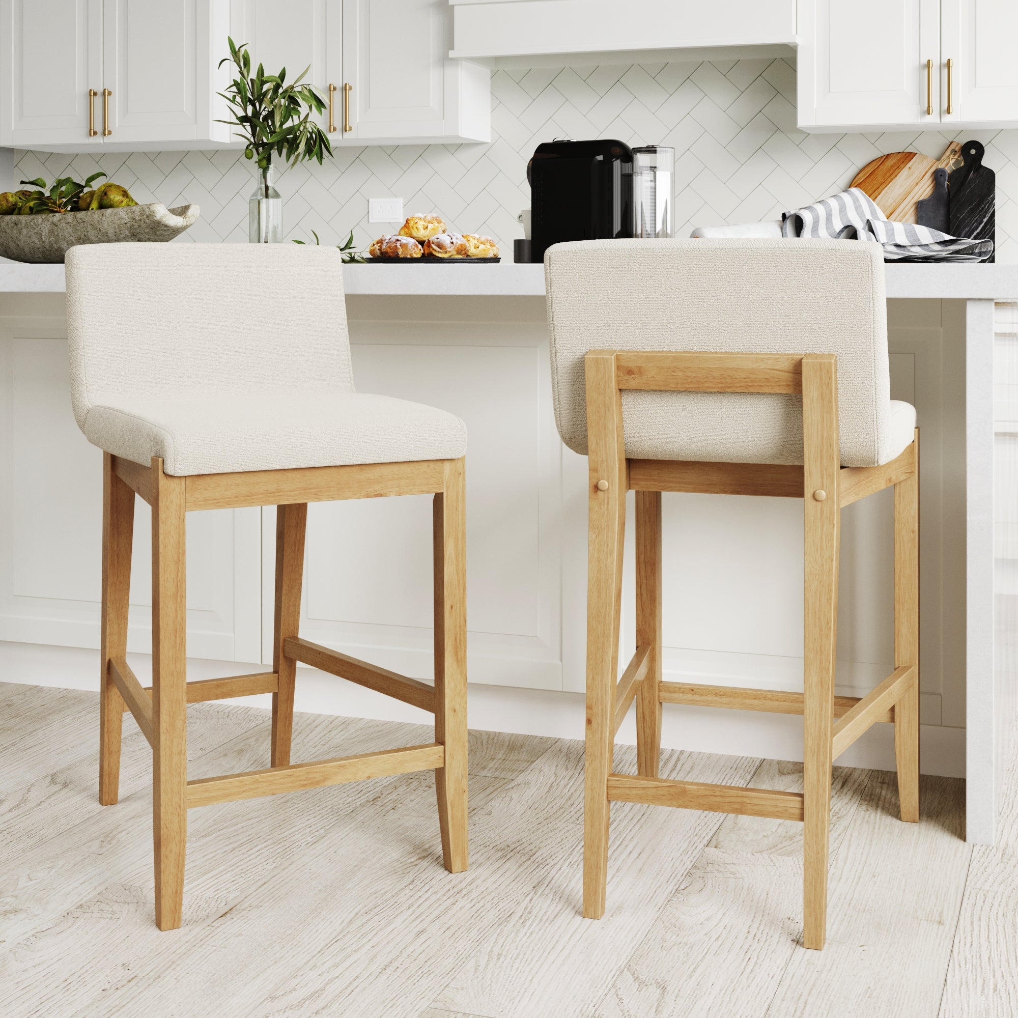 Two wooden bar stools with light beige upholstered seats are placed at a white kitchen counter. The kitchen features white cabinets, a herringbone backsplash, and various items on the counter.