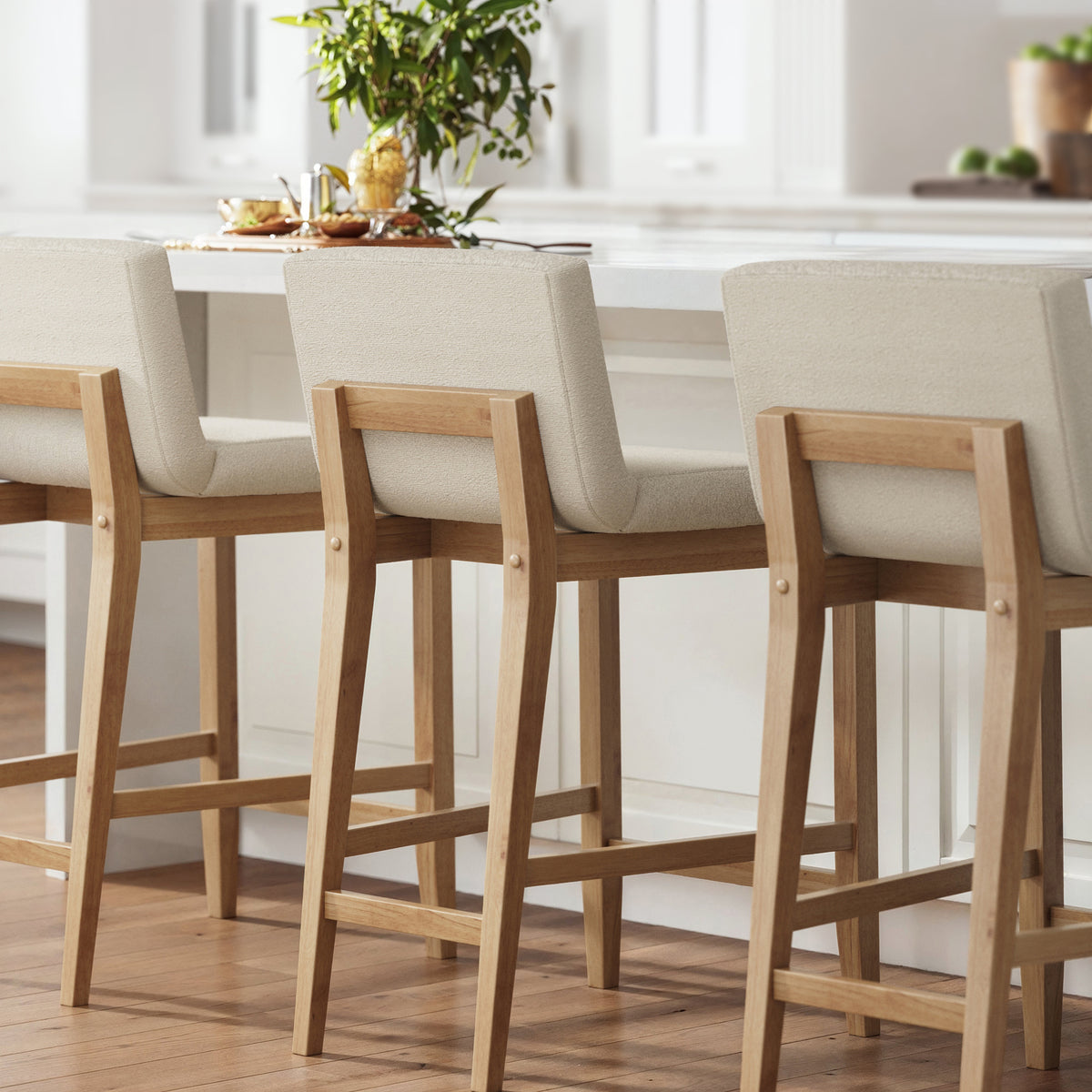 Three light-colored upholstered bar stools with wooden frames lined up at a kitchen island.
