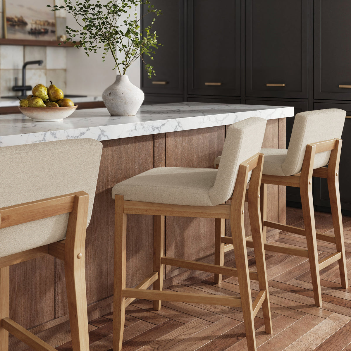 Three modern wooden bar stools with upholstered beige seats arranged along a kitchen island with a marble countertop.