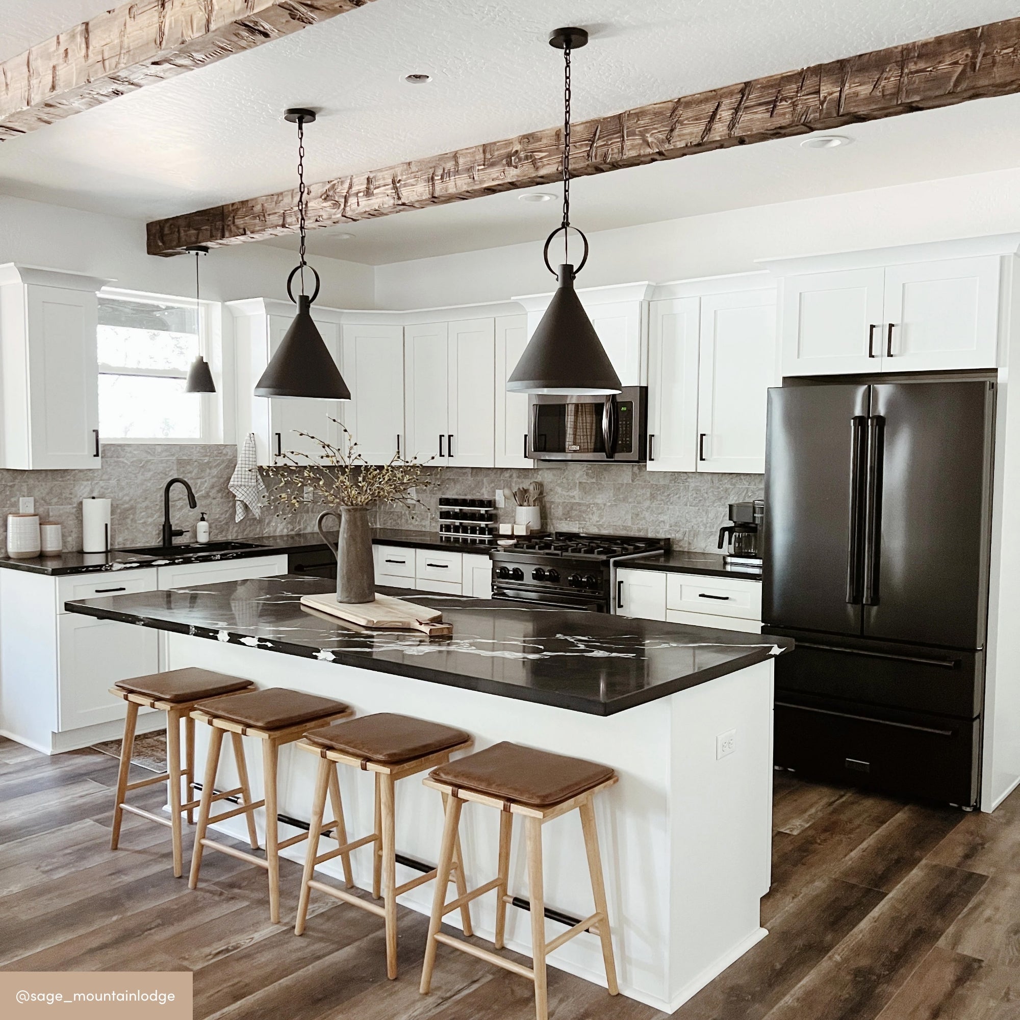 Modern kitchen with white cabinets, black appliances, a large island with black countertop, four wooden stools, pendant lights, wooden ceiling beams, and a vase with branches on the island. Wood flooring completes the space.