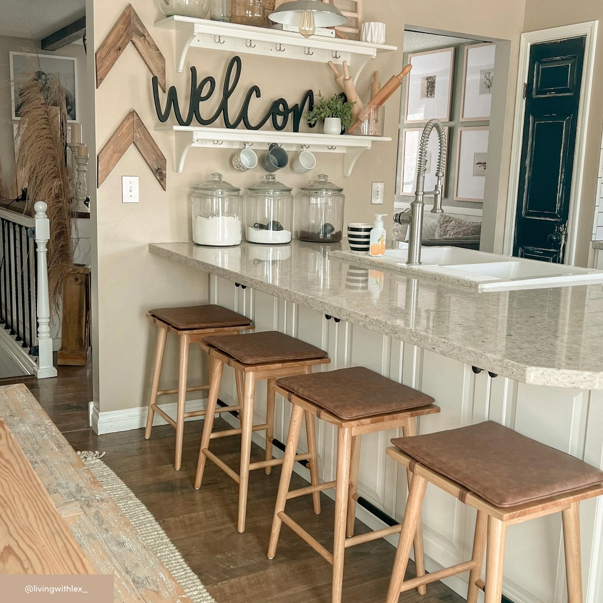 A cozy kitchen with a white countertop, four wooden stools, glass jars, mugs hanging under a shelf, and a large black welcome sign on the wall. Decor includes greenery and rustic accents.