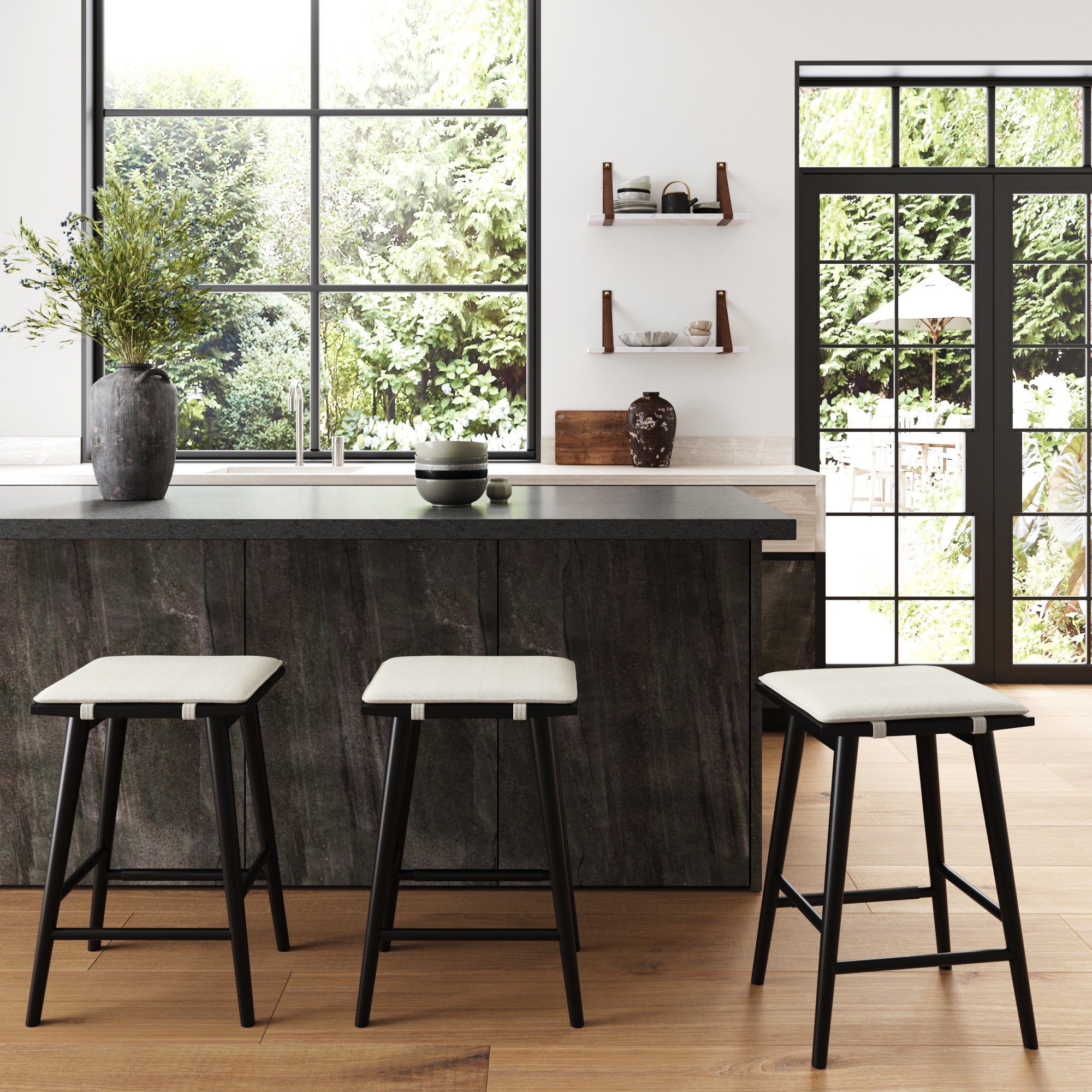 Modern kitchen with large windows, a dark island countertop, three cushioned bar stools, wooden floors, decorative vases, shelves with bowls, and a view of a green garden outside.