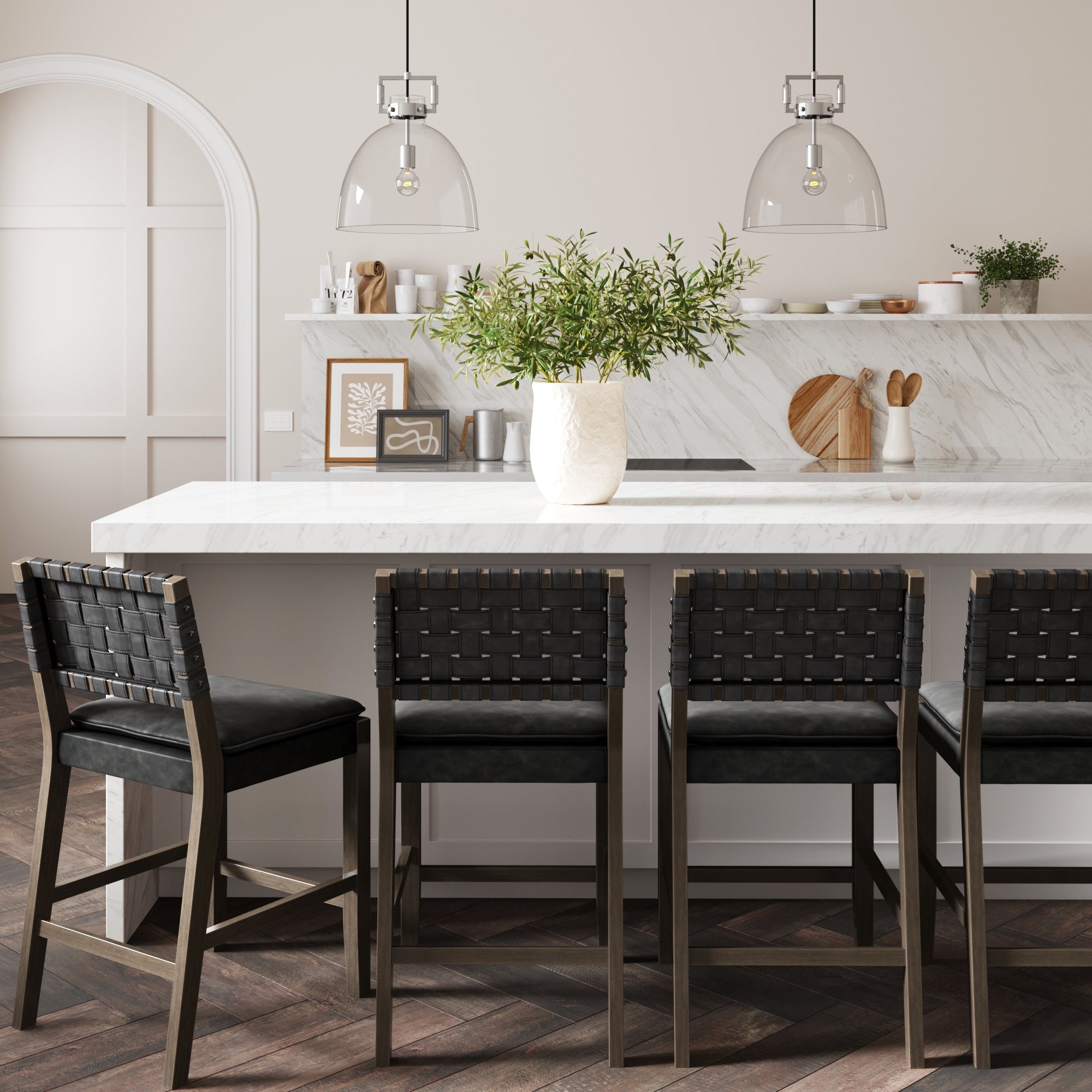 Modern kitchen with a white marble island, four black woven barstools, a large vase with greenery, and pendant lights hanging above. Neutral decor with wood and marble accents in the background.