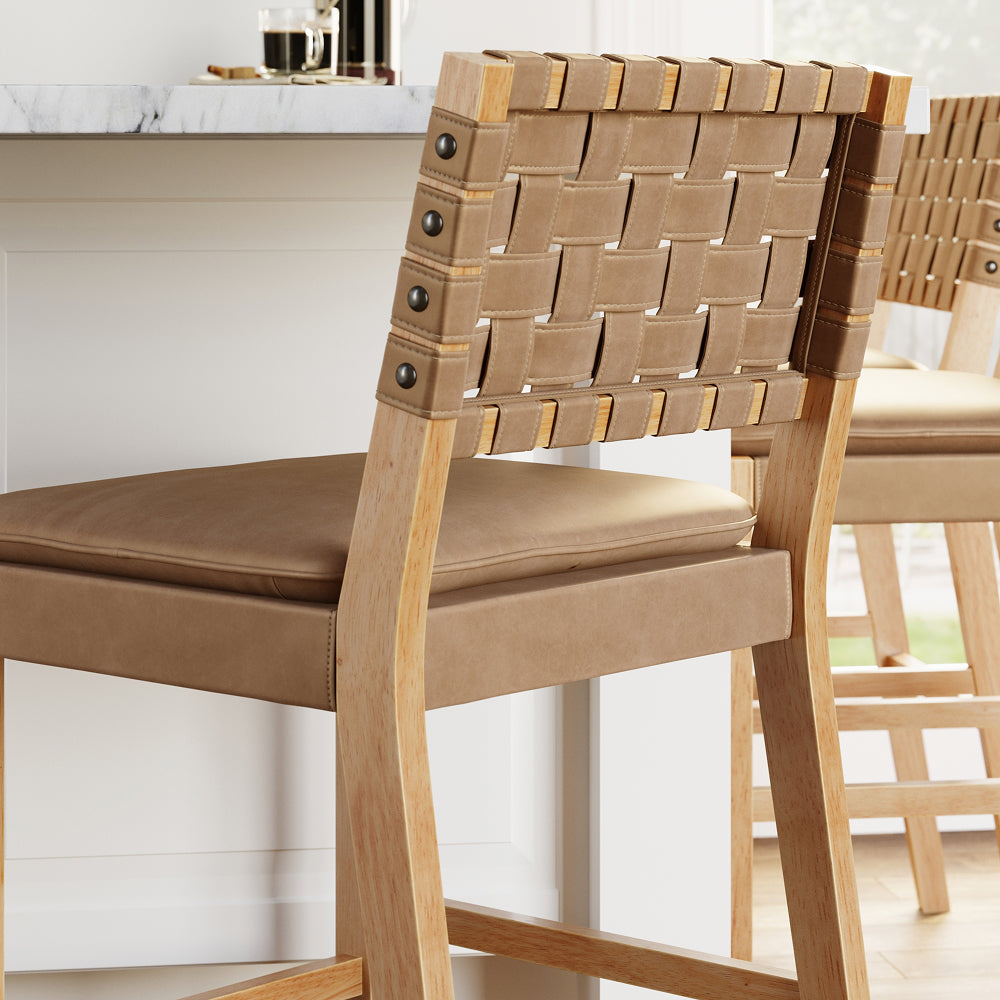 Light wood bar stool with a tan upholstered seat and a backrest featuring a woven strap design attached with decorative studs.
