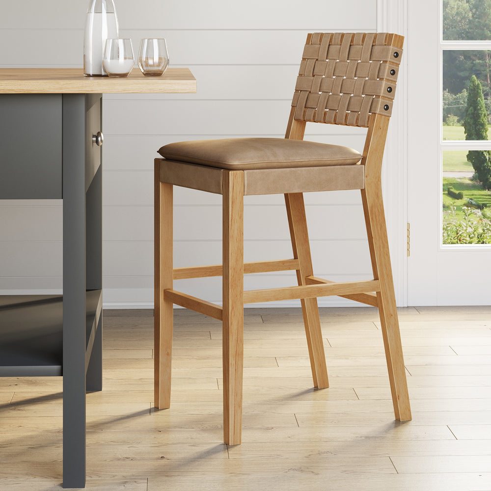 A wooden bar stool with a tan upholstered seat and a woven strap backrest, positioned next to a kitchen island.