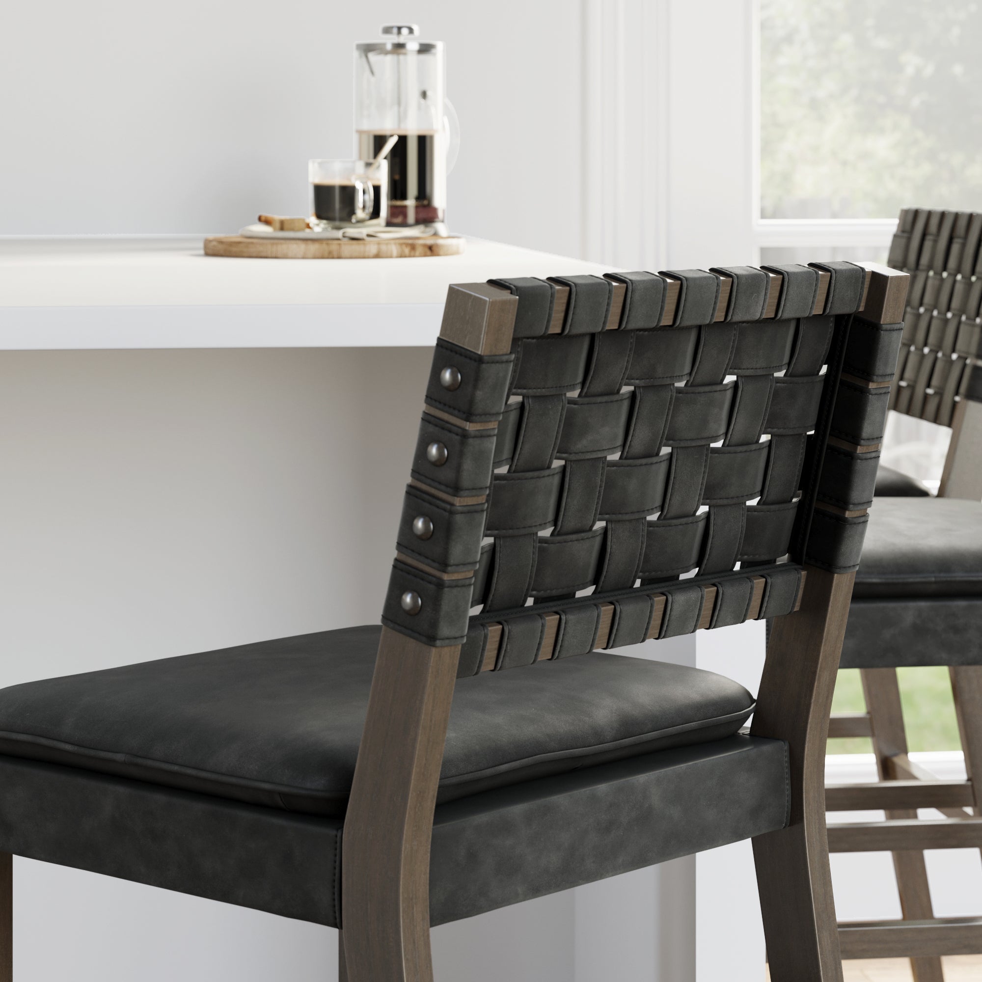 A close-up of a bar stool with a woven black leather backrest and a cushioned seat, beside a white counter. On the counter are a French press, a glass of coffee, and a small wooden tray. Light streams in through a window.