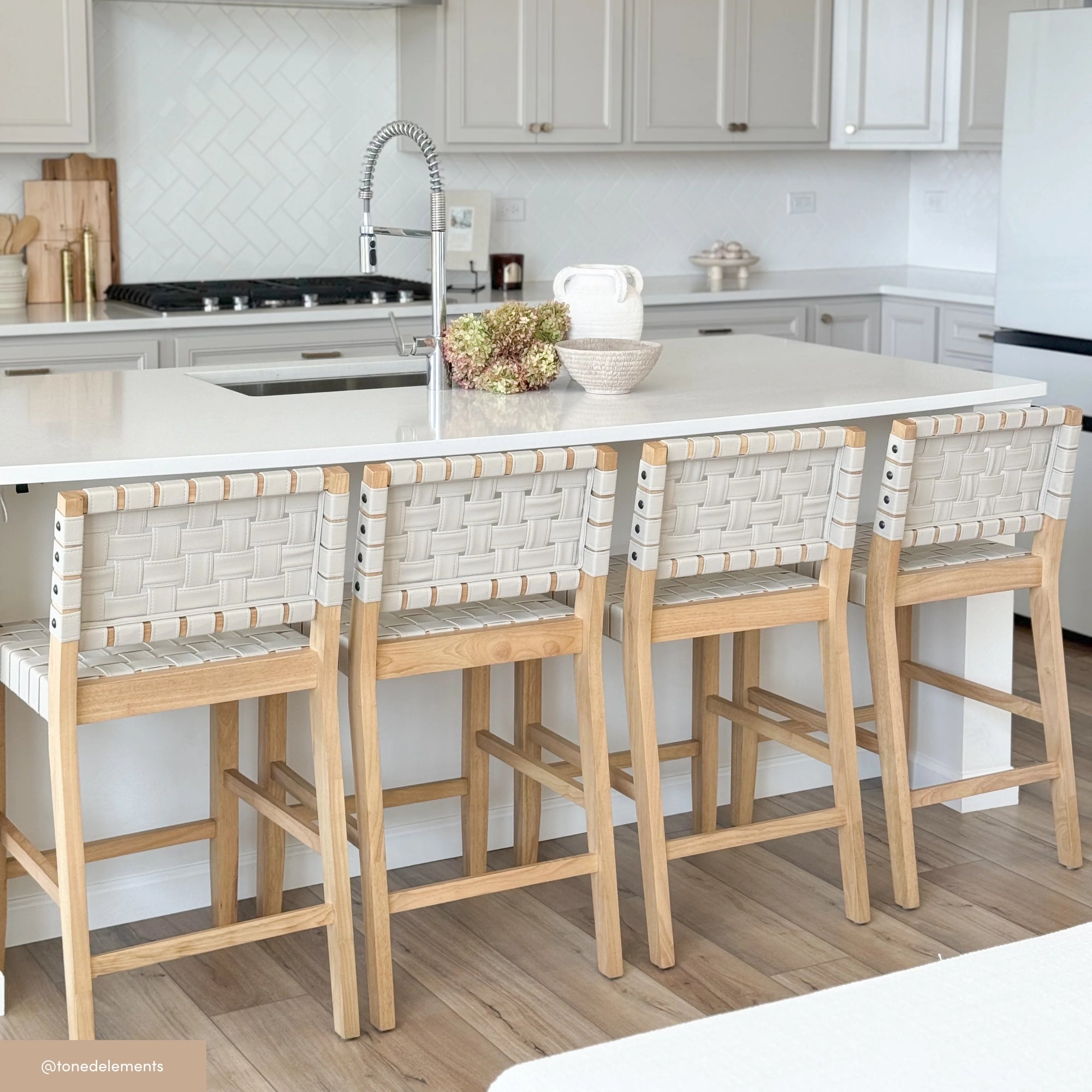 A modern kitchen with a white island, four wooden bar stools with woven seats, a sink with a tall faucet, and light-colored cabinets and flooring. A decorative bowl and a bouquet are on the counter.