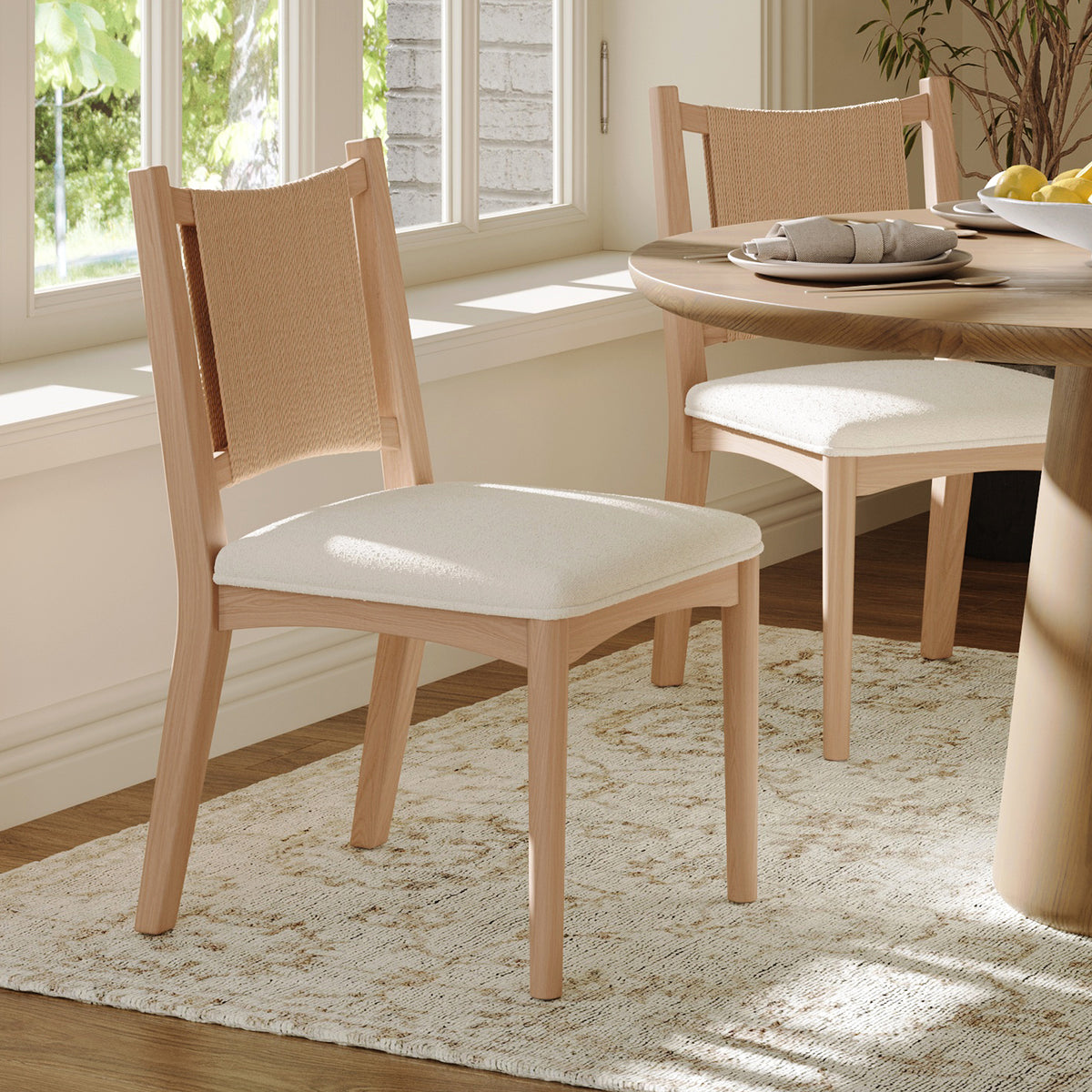 Two light wood dining chairs with cream upholstered seats and woven backrests positioned at a round wooden table.