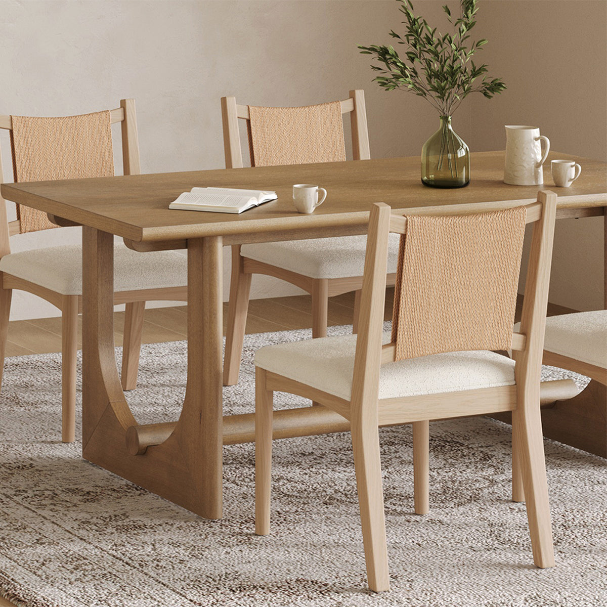 A wooden dining table with four chairs featuring woven backrests and upholstered seats, set on a patterned rug with a book, mugs, and a vase on the table.