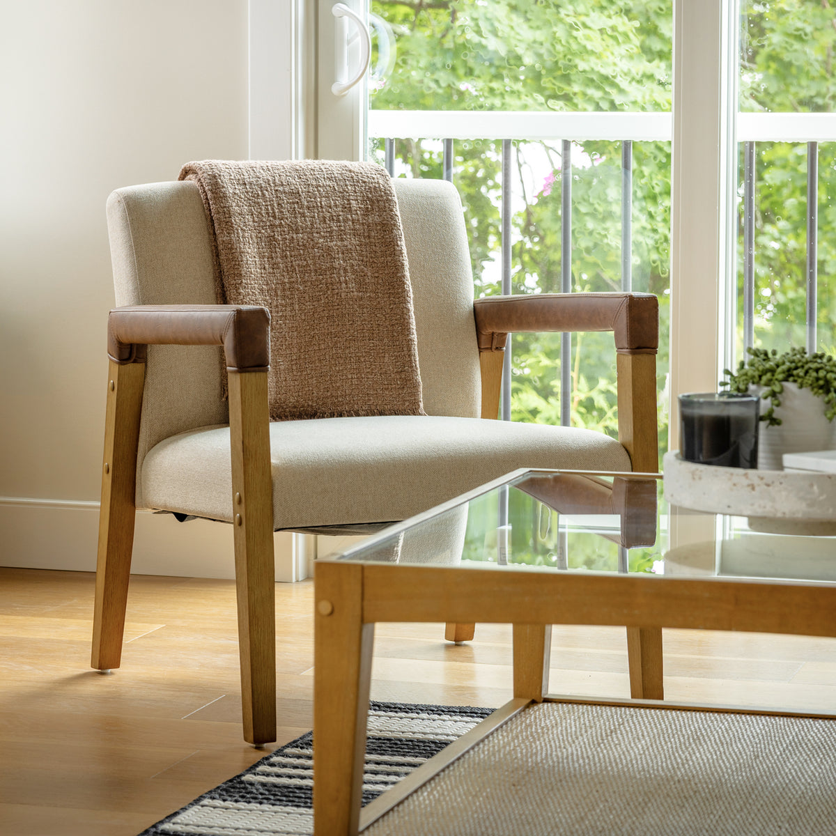 Nathan James Wood & Faux Leather Upholstered Accent Chair with light-colored upholstery, a solid wood frame, brown faux leather armrest covers, and a throw blanket draped over the back.