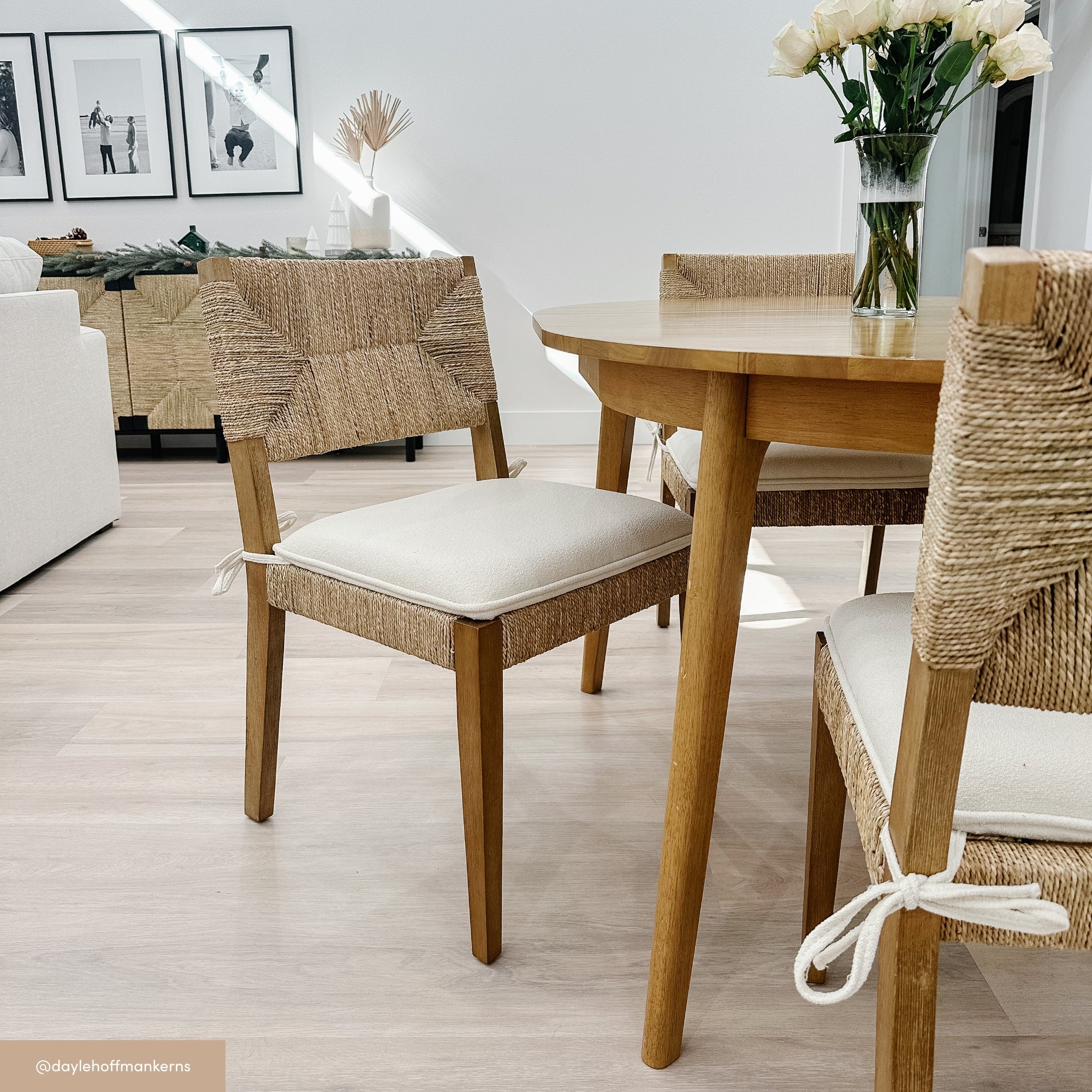 A light-filled dining area with wooden chairs featuring woven backs and white cushions around a round wooden table topped with a glass vase of white roses. Art and decor are visible in the background.