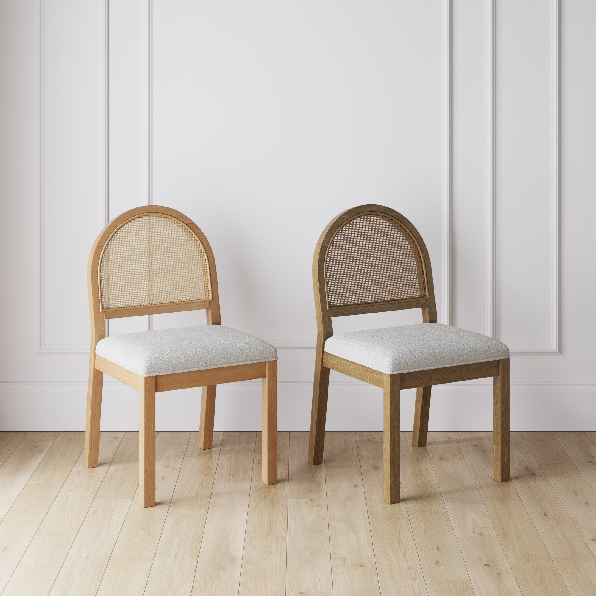 Two wooden chairs with rounded backs and light gray seat cushions are placed side by side on a light wooden floor in front of a white paneled wall. The chairs have different wood finishes.