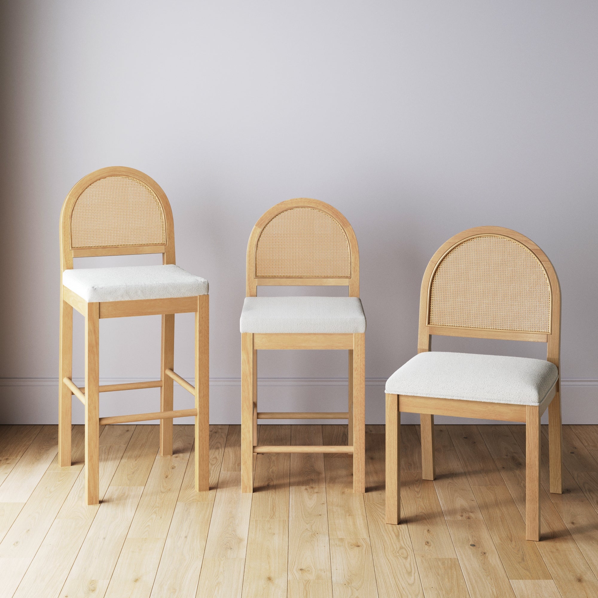 Three wooden chairs with light-colored cushioned seats and rounded rattan backs are arranged in a row on a wooden floor against a plain light-colored wall. Each chair is a different height.