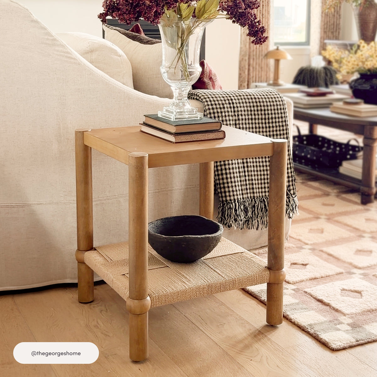 Nathan James Coastal Wood & Seagrass 2-Tier Side Table Light Brown with a top shelf holding books and a vase of flowers, and a lower shelf featuring a faux seagrass base with a dark decorative bowl.