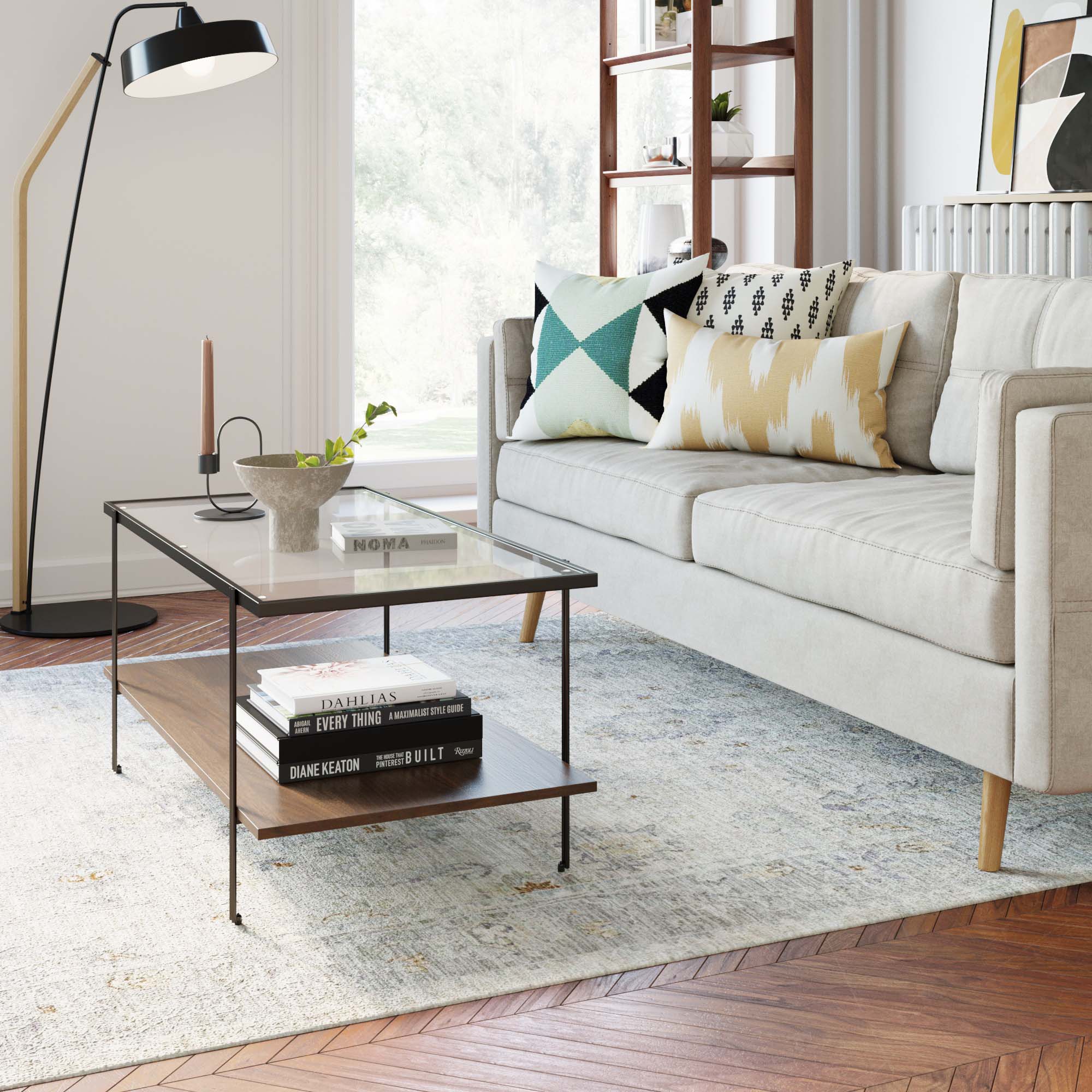 Bright living room with a beige sofa, geometric cushions, a glass coffee table holding a plant and candle, books underneath, a standing lamp, and a patterned rug on a wooden floor. Large windows let in natural light.