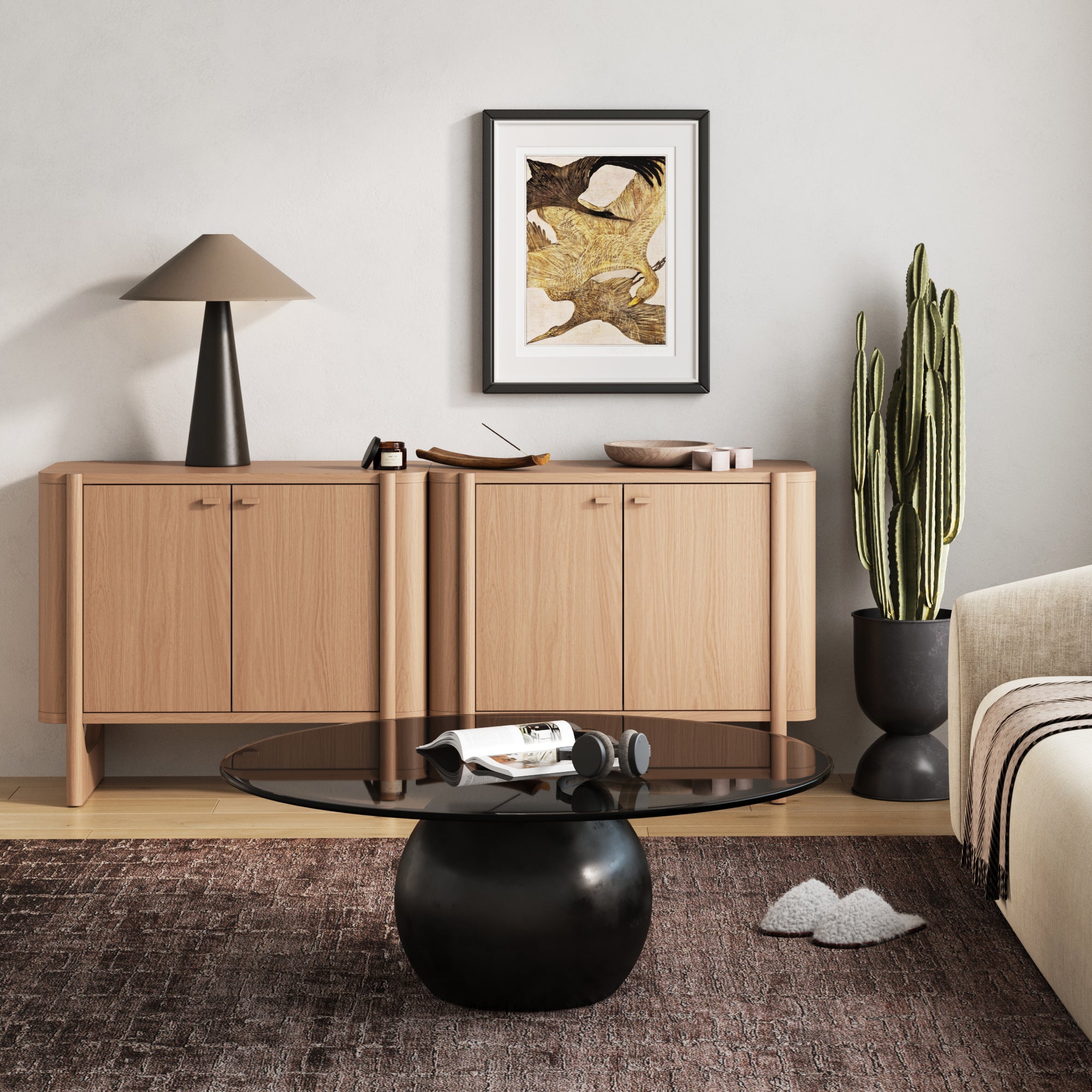 A modern living room with a light wood sideboard, a black lamp, a tall cactus in a planter, abstract wall art, a round glass coffee table, and a beige sofa with a brown striped throw on a patterned rug.
