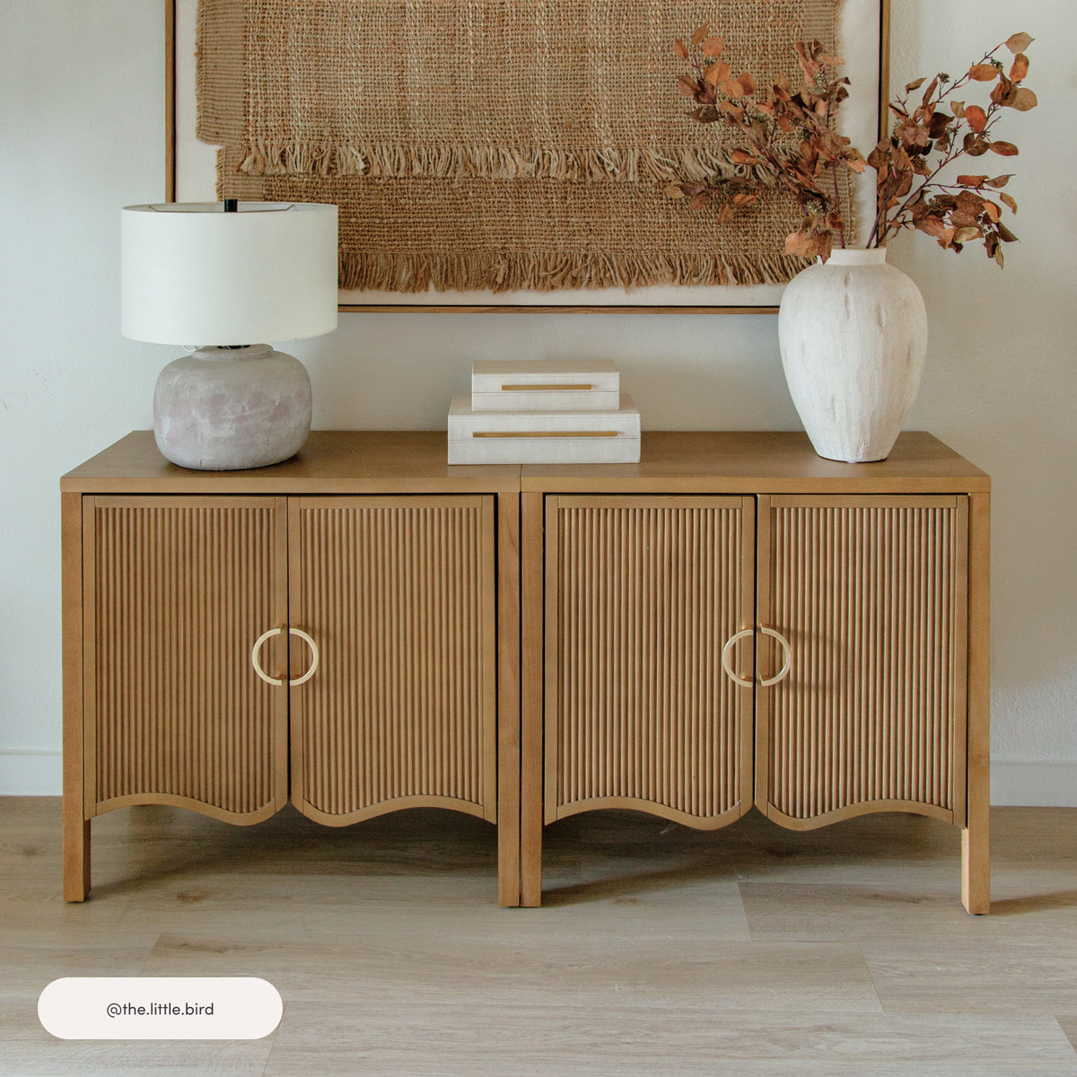 Nathan James Wood Transitional Fluted Cabinet w/ Brass Handle Light Brown featuring four fluted doors and circular brass handles, topped with a table lamp, a stack of two boxes, and a vase of dried branches.