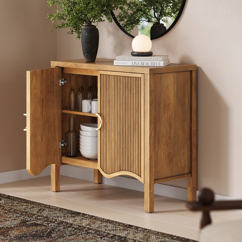 A wooden sideboard with one open door, revealing glasses and stacked bowls inside, with a lamp and books on the surface.