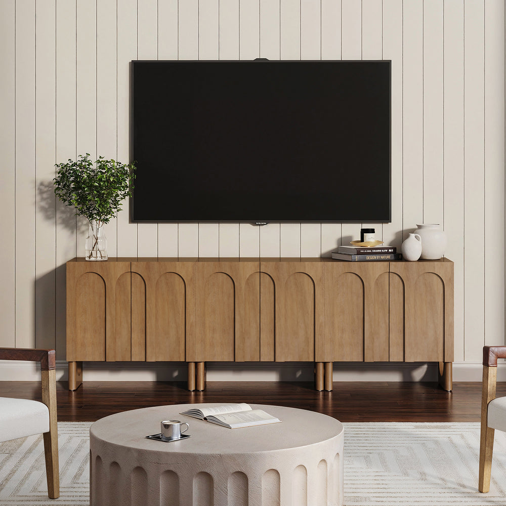 A large flat-screen television mounted above a wood console table with an arched door design, featuring a small potted plant, two vases, and a stack of books on top.