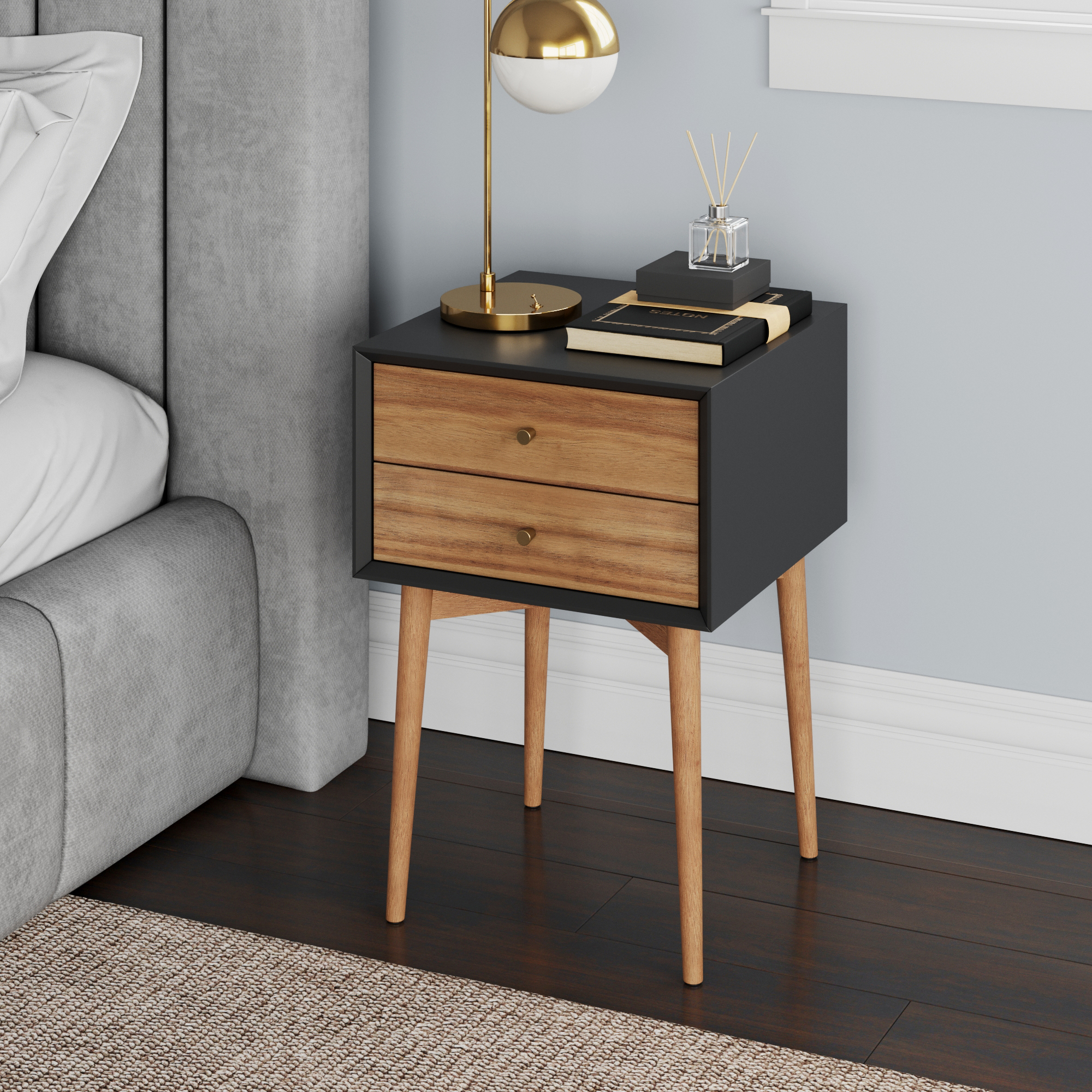 A modern nightstand with two wooden drawers and tapered legs stands beside a bed. On top are a gold and white lamp, books, and decorative items. The room has dark wood floors and a light wall.