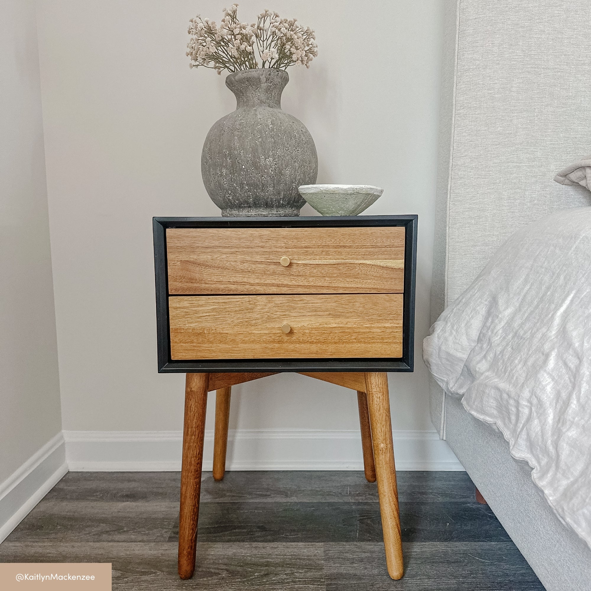 A wooden bedside table with two drawers stands beside a bed. On top, there is a gray textured vase with dried flowers and a small ceramic bowl. The floor is dark wood and the wall is white.