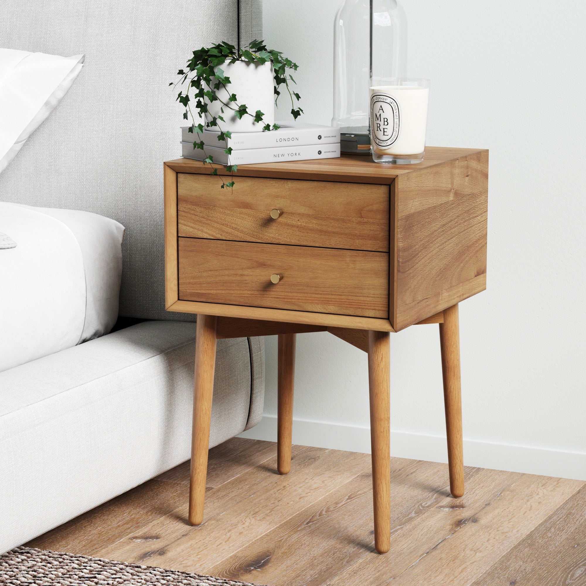 A wooden nightstand with two drawers and angled legs stands next to a bed. On top are a trailing potted plant, two books, a clear glass lamp, and a white candle. The floor is wood and the wall is light-colored.